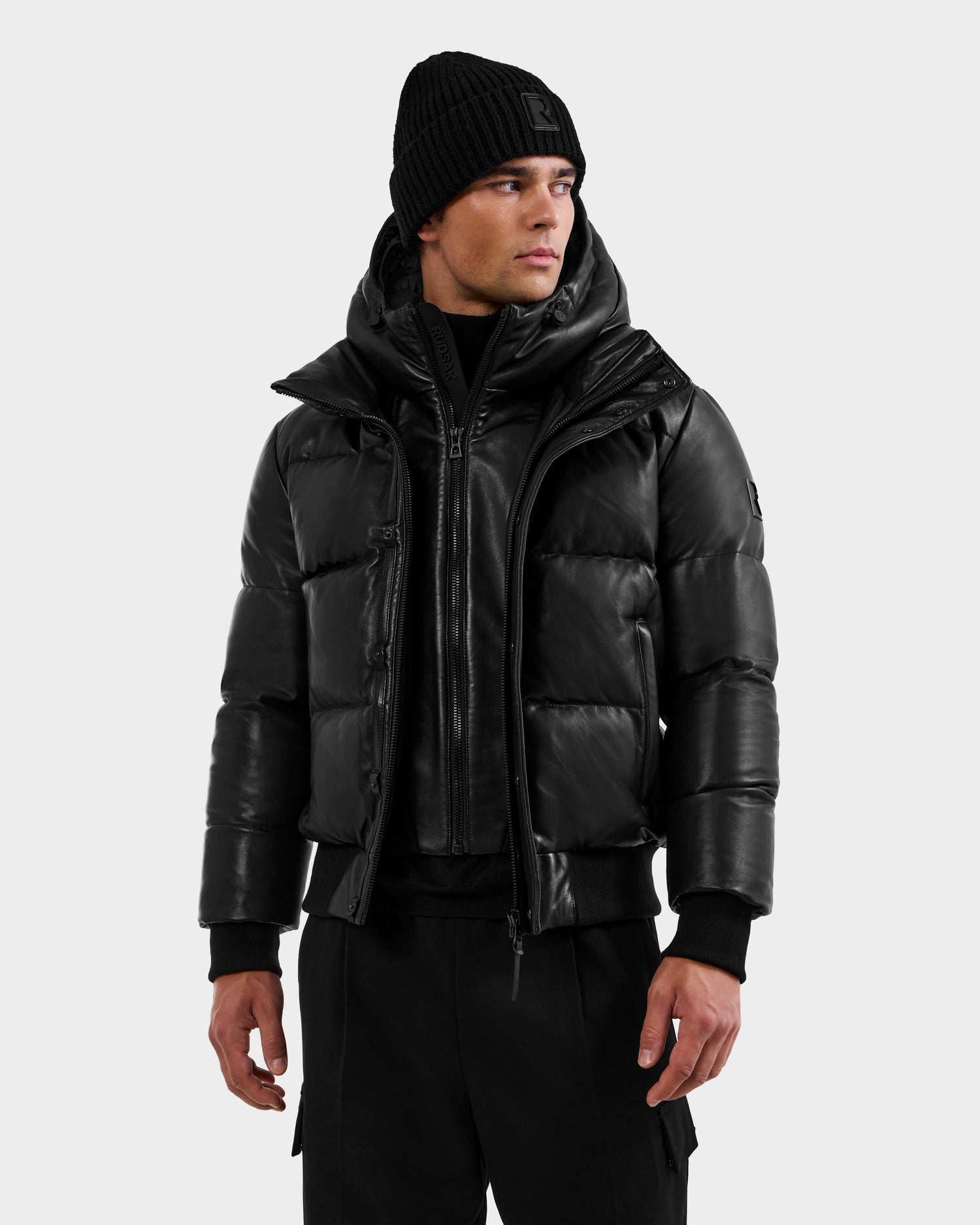 A man wears the RUDSAK BOND X - 6122525 BLACK puffer jacket, black beanie, sweater, and pants, standing against a plain light gray background and looking slightly to his left.