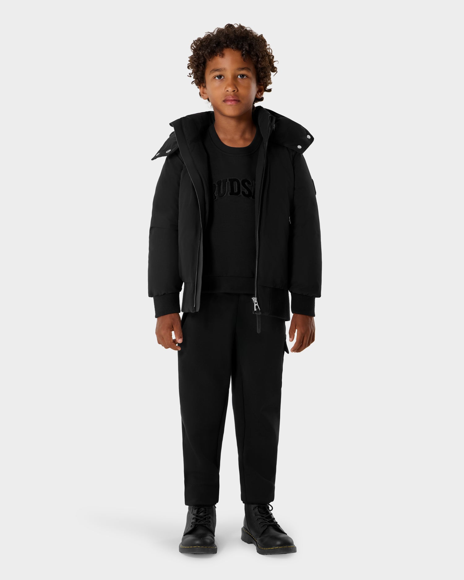A young boy with curly hair faces forward, wearing the Rudsak BOOKER K - 0124500 Black jacket, black sweatshirt, pants, and boots against a plain light gray background.