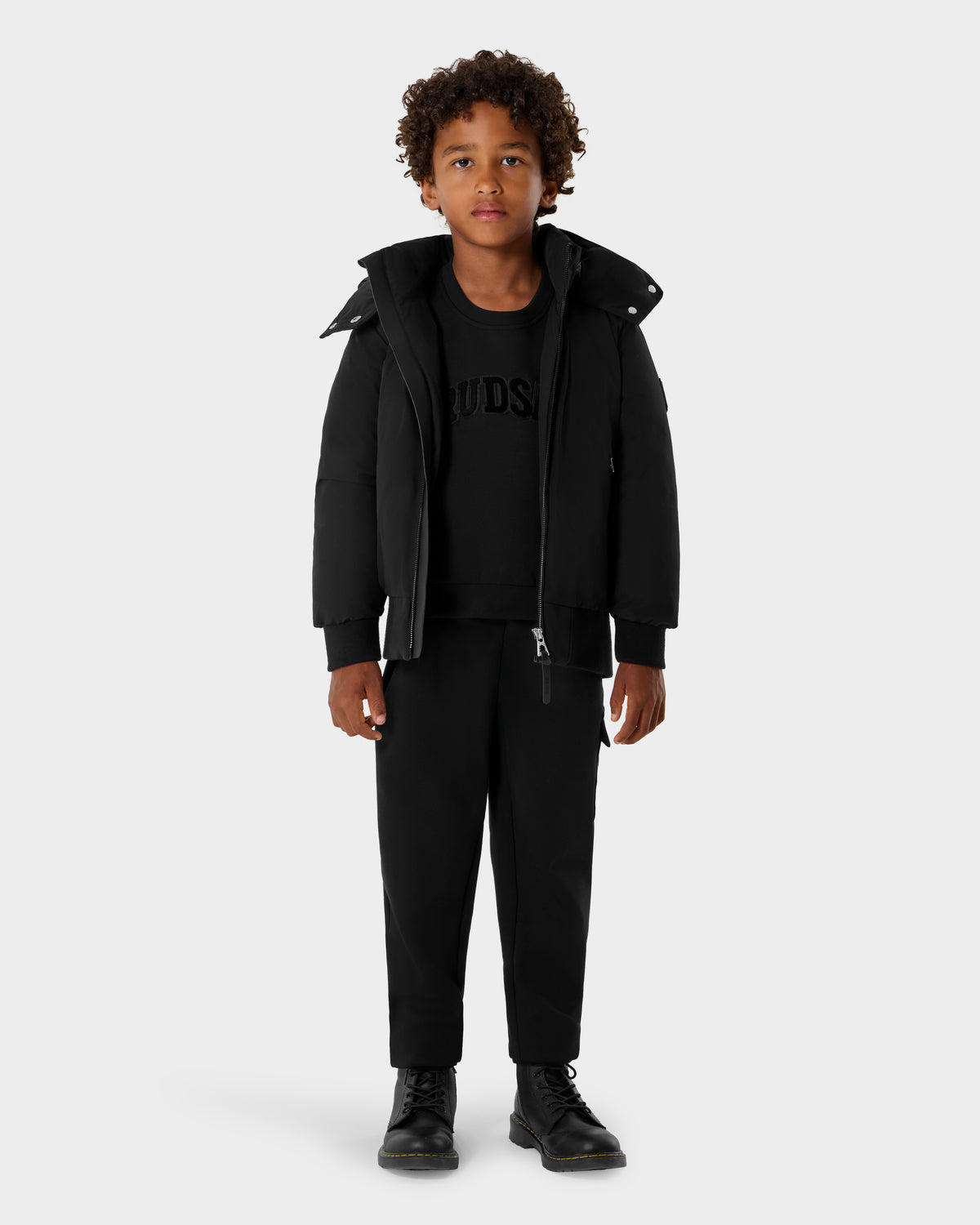 A young boy with curly hair faces forward, wearing the Rudsak BOOKER K - 0124500 Black jacket, black sweatshirt, pants, and boots against a plain light gray background.
