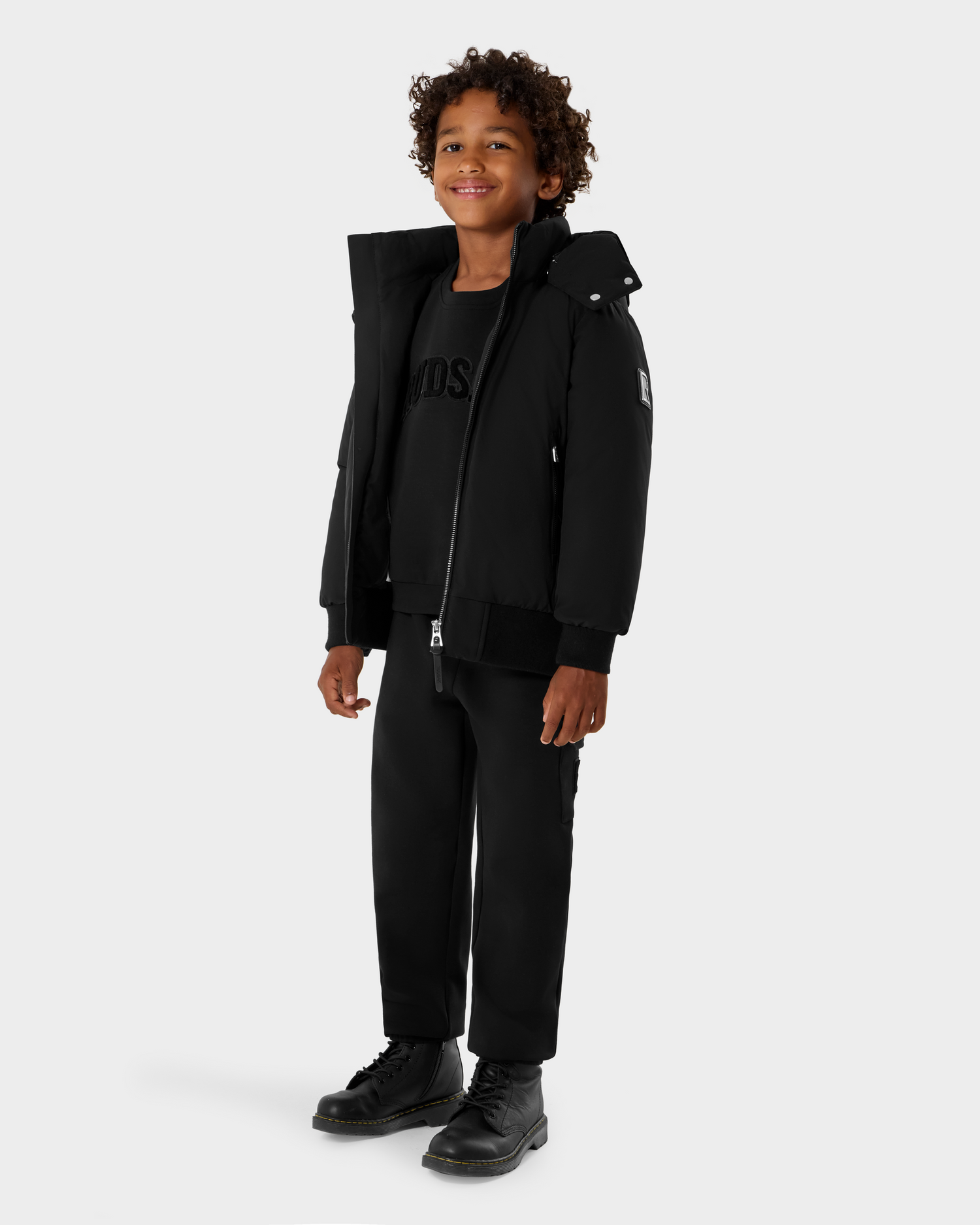 A smiling curly-haired child stands against a light gray background wearing the Rudsak BOOKER K - 0124500 Black bomber jacket, paired with black sweatshirt, pants, and boots.