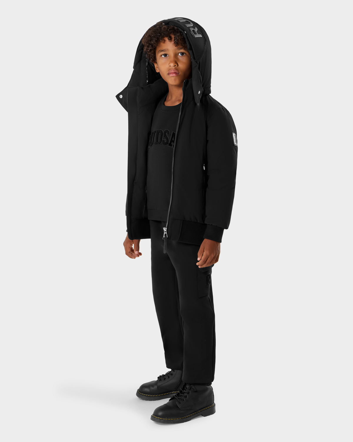 A young boy with curly hair wears the Rudsak BOOKER K - 0124500 Black kids’ quilted puffer jacket, paired with a black shirt, pants, and boots, standing against a plain light gray background.