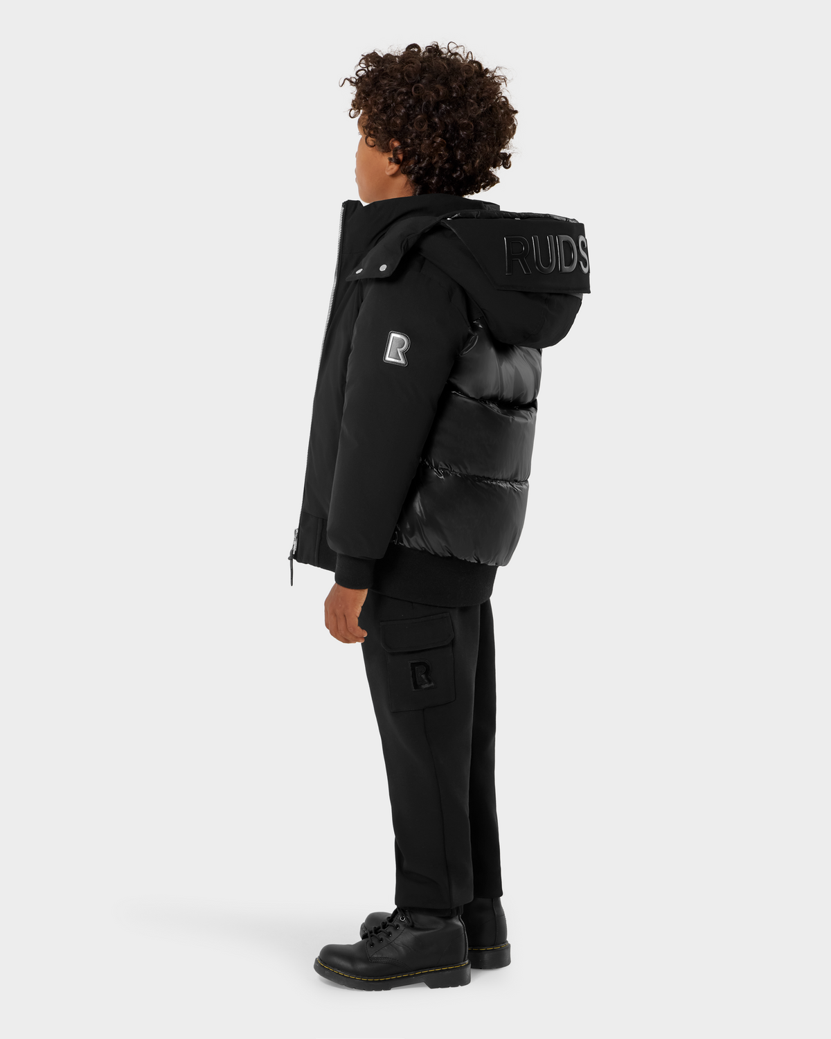 A person with curly hair wears the Rudsak BOOKER K - 0124500 Black weather-resistant puffer jacket, black pants, and black shoes, standing against a plain white background and facing left.