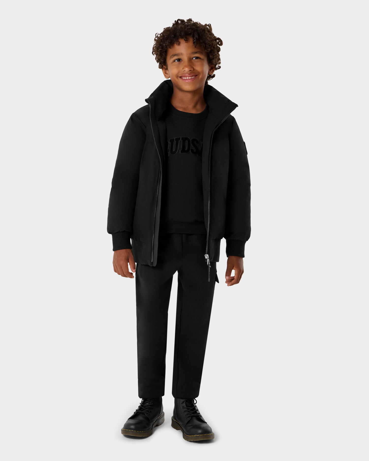 A young boy with curly hair wears Rudsak’s BOOKER K - 0124500 Black bomber jacket, paired with a black sweatshirt, pants, and boots while standing and smiling against a light gray background.