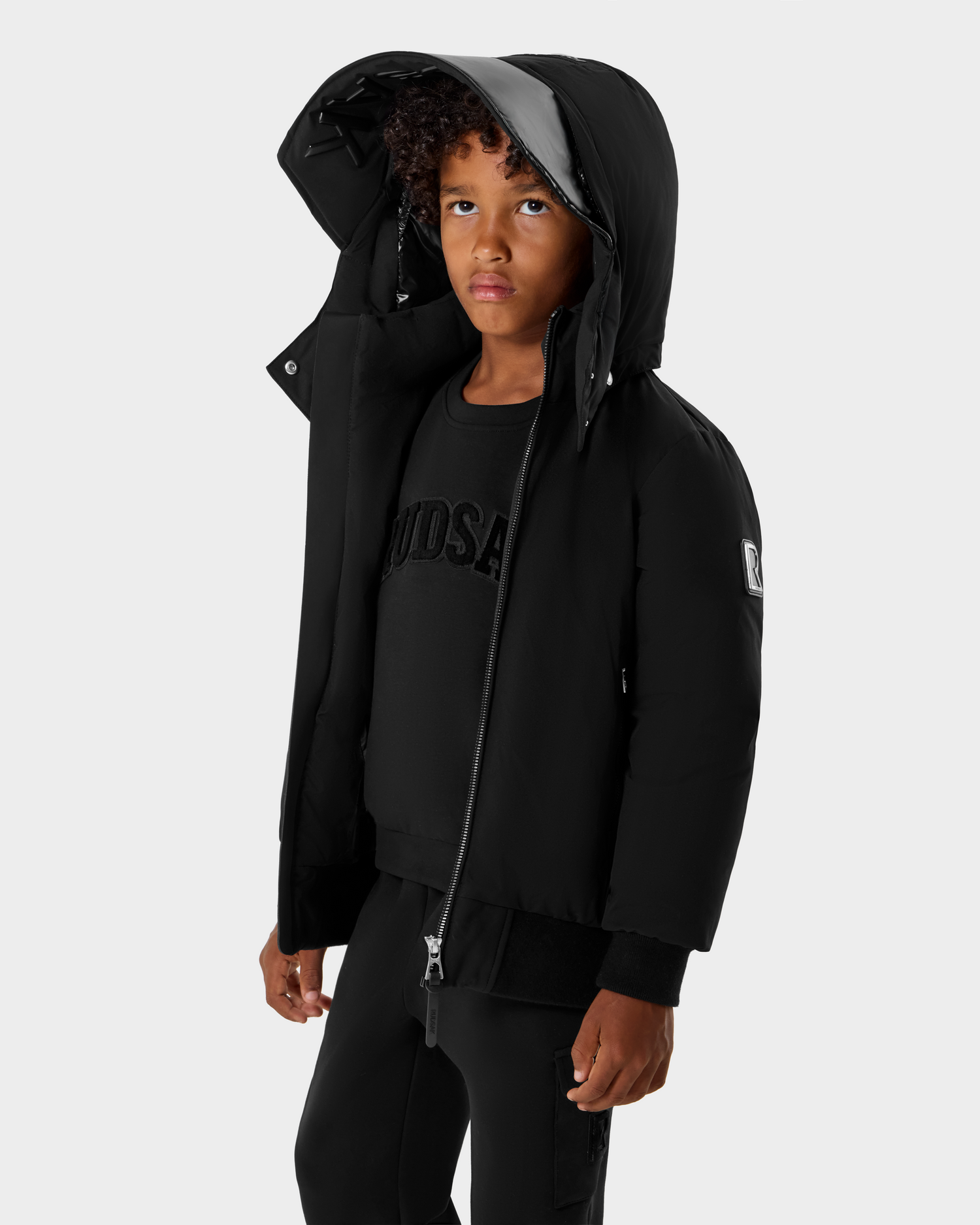 A young child with curly hair wears the Rudsak BOOKER K - 0124500 Black kids' quilted puffer jacket over black clothes, standing against a plain light gray background and looking slightly upward.