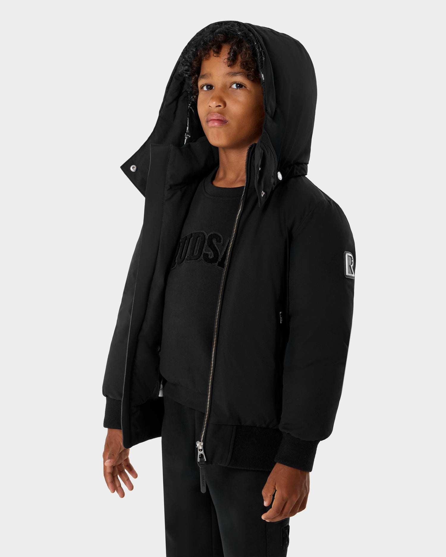 A young child with curly hair models the Rudsak BOOKER K - 0124500 Black kids' quilted puffer jacket with a removable hood over a black shirt and pants, standing against a light gray background. The weather-resistant jacket is unzipped.