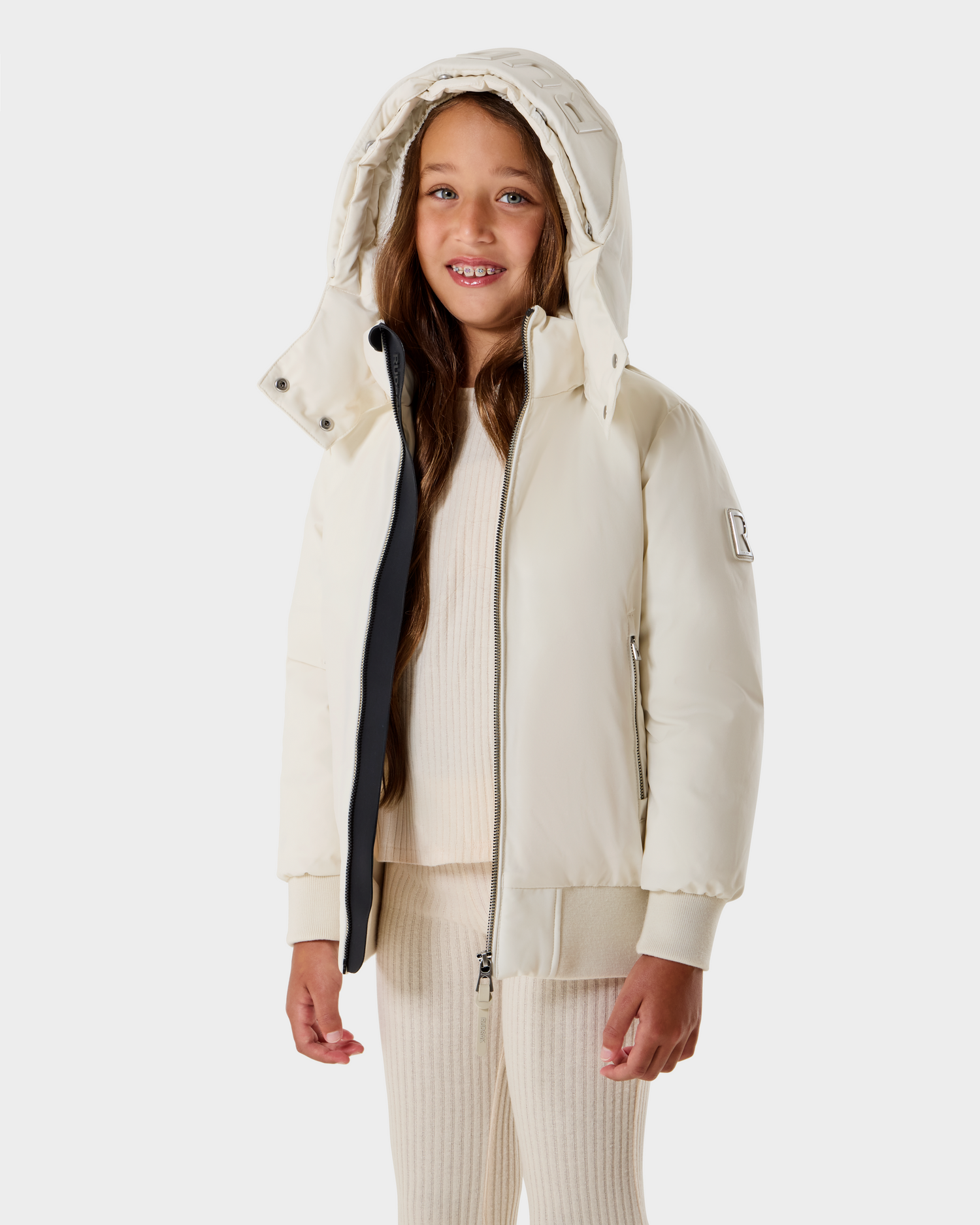 A young girl with long brown hair smiles while wearing the Rudsak BOOKER K - 0124500 Vanilla kids' quilted puffer jacket, paired with matching ribbed knit pants and top, standing against a plain light background.