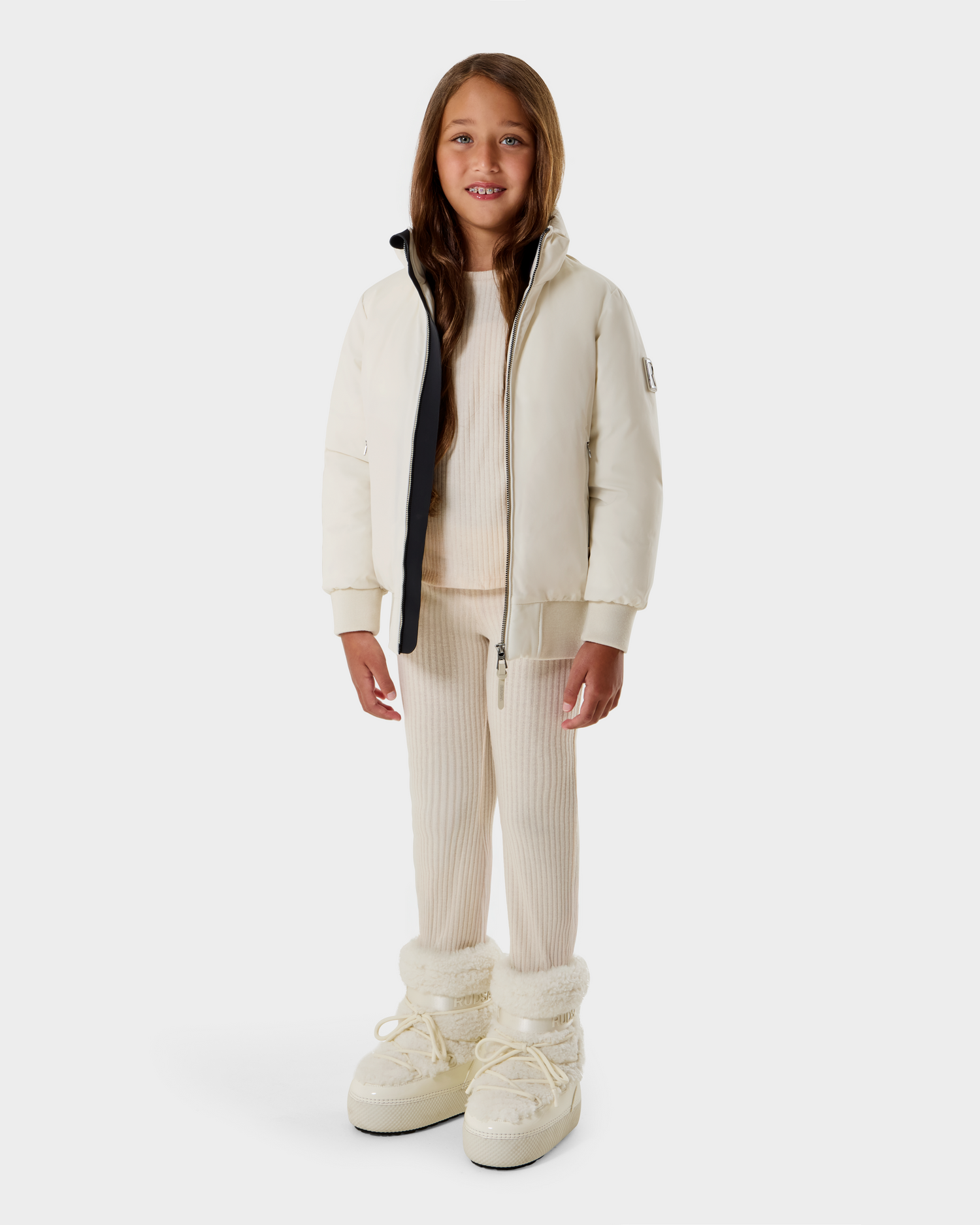 A young girl with long brown hair smiles while wearing the Rudsak BOOKER K - 0124500 Vanilla kids' quilted puffer jacket, a cream ribbed sweater, matching pants, and fluffy white boots against a plain light gray background.