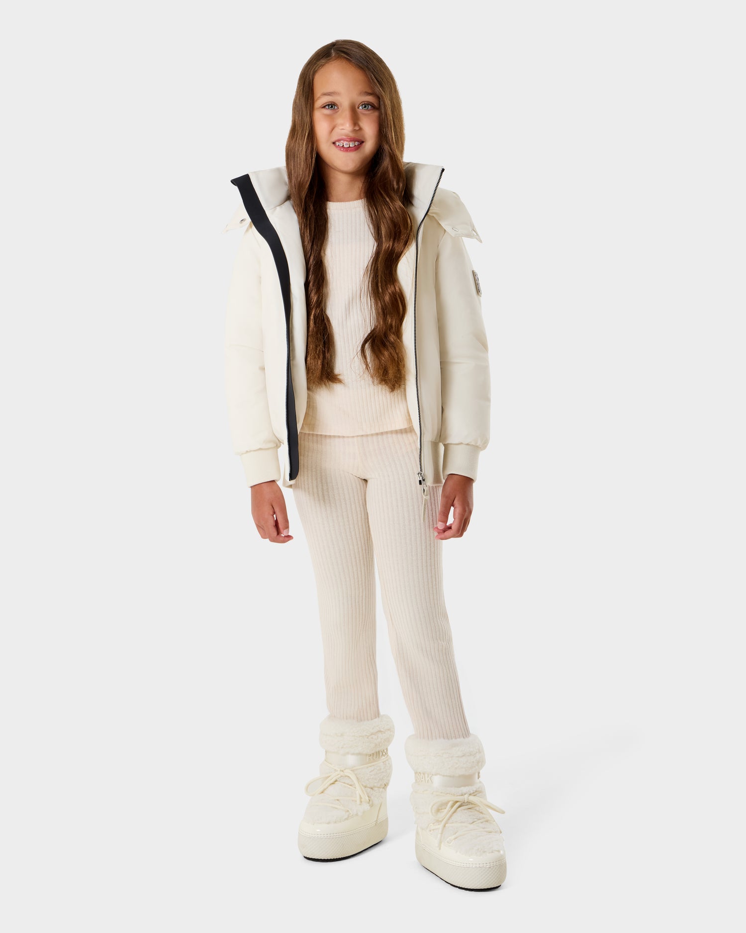 A young girl with long brown hair wears the Rudsak BOOKER K - 0124500 Vanilla kids' quilted puffer jacket, complemented by a matching sweater, ribbed leggings, and white winter boots as she stands smiling against a plain light background.