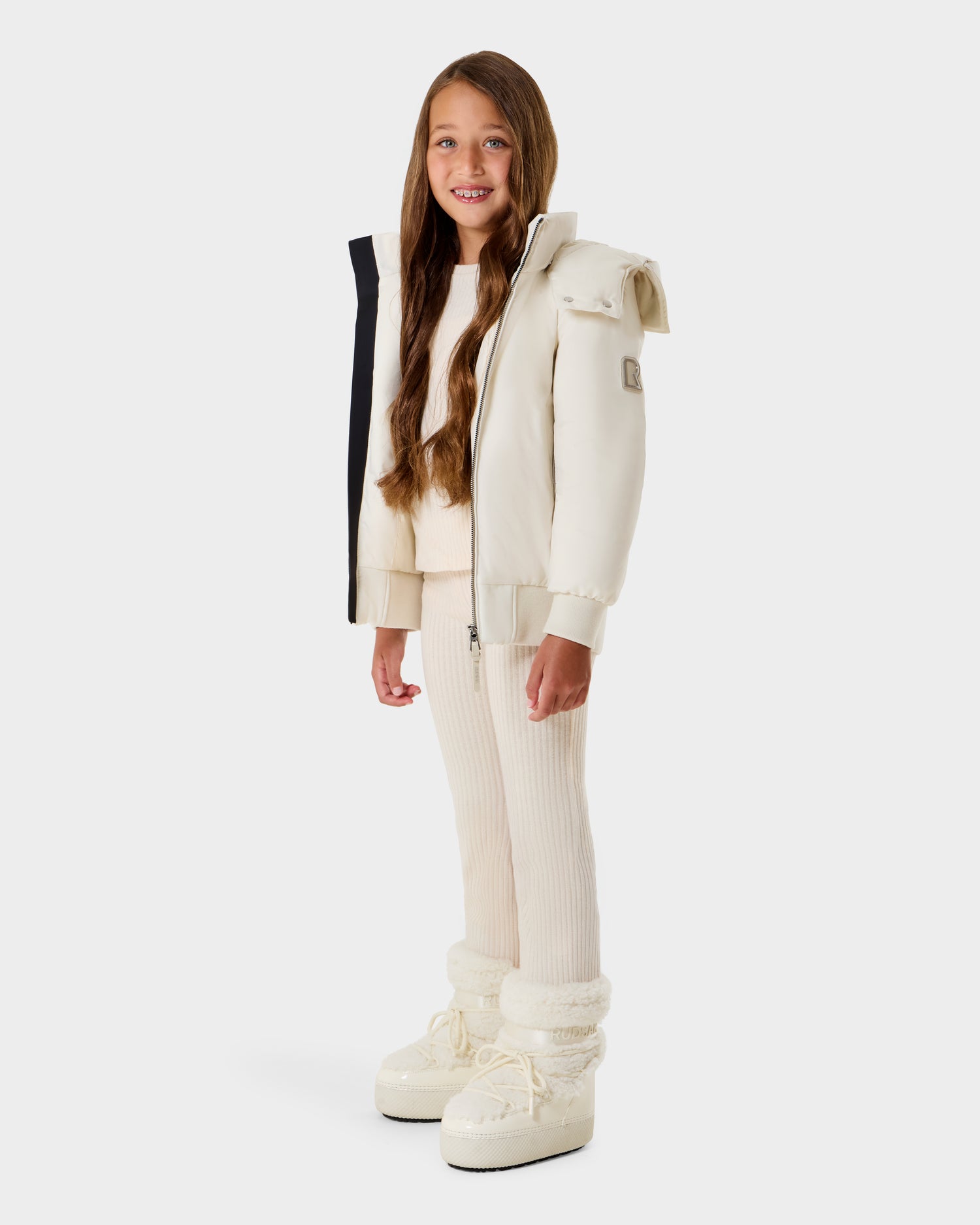 A young girl with long brown hair smiles, wearing the Rudsak BOOKER K - 0124500 Vanilla kids' quilted puffer jacket with matching ribbed pants and winter boots, standing against a plain white background.