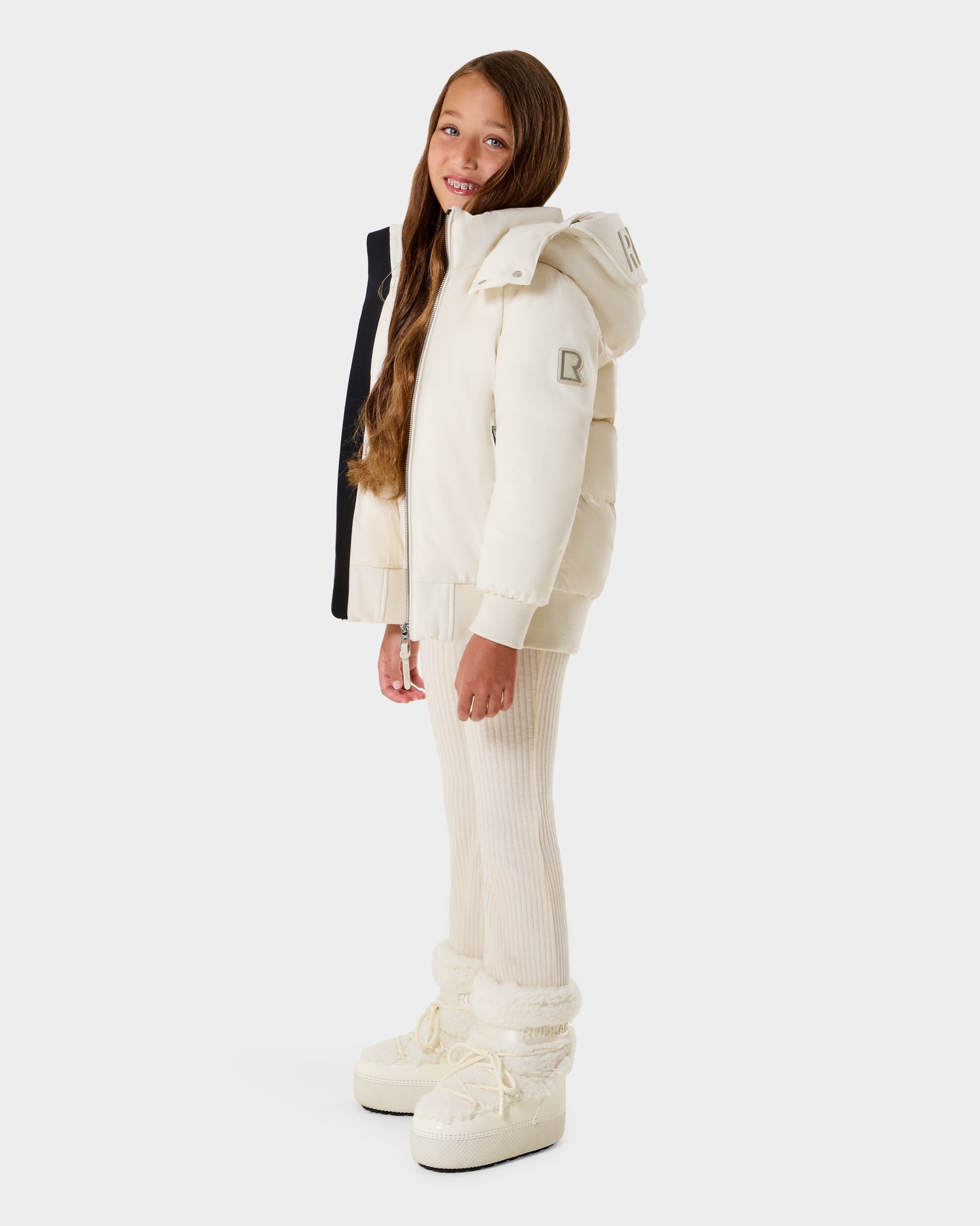 A young girl smiles at the camera, wearing the Rudsak BOOKER K - 0124500 Vanilla kids' quilted puffer jacket with a removable hood, ribbed pants, and white snow boots. She has long brown hair and stands against a plain white background.