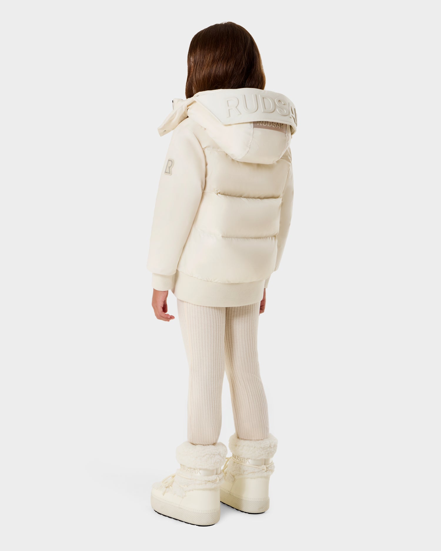 A young child with long brown hair, seen from behind, wears the Rudsak BOOKER K - 0124500 Vanilla kids' quilted puffer jacket with branded hood, cream leggings, and matching boots against a plain light background.
