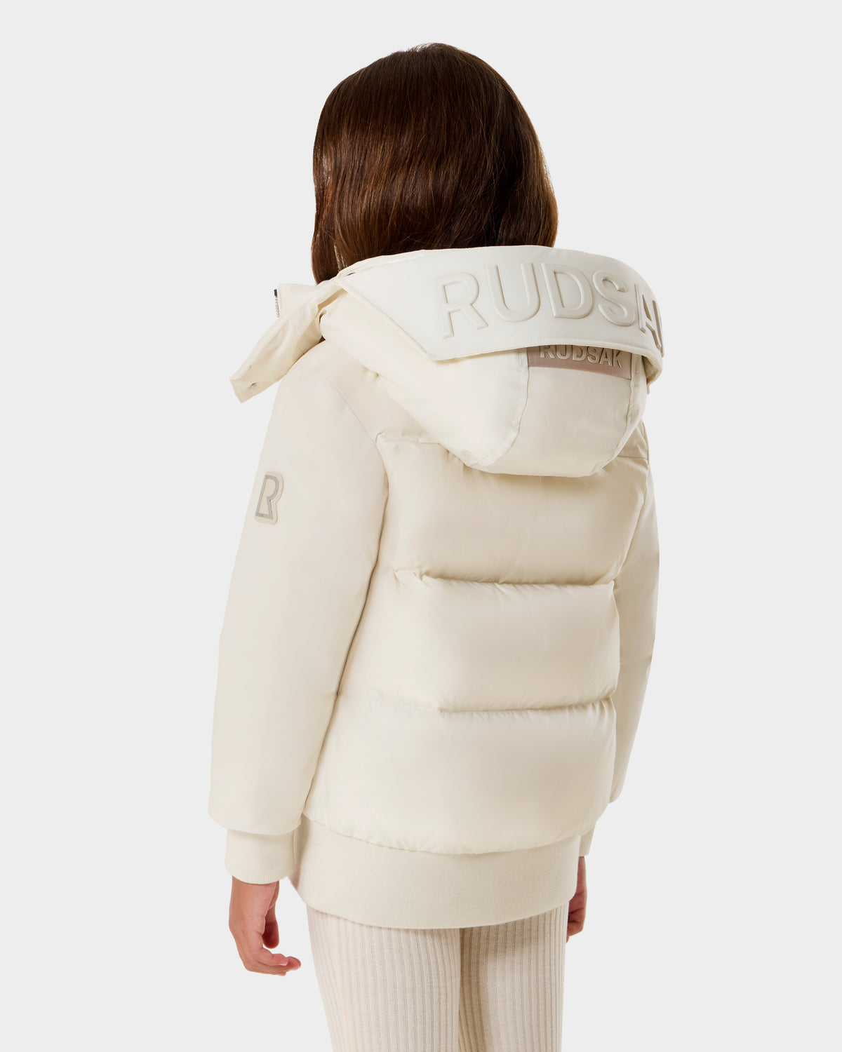 A child with long brown hair wears the Rudsak BOOKER K - 0124500 Vanilla kids' quilted puffer jacket with a removable hood, standing against a plain white background and facing away from the camera.