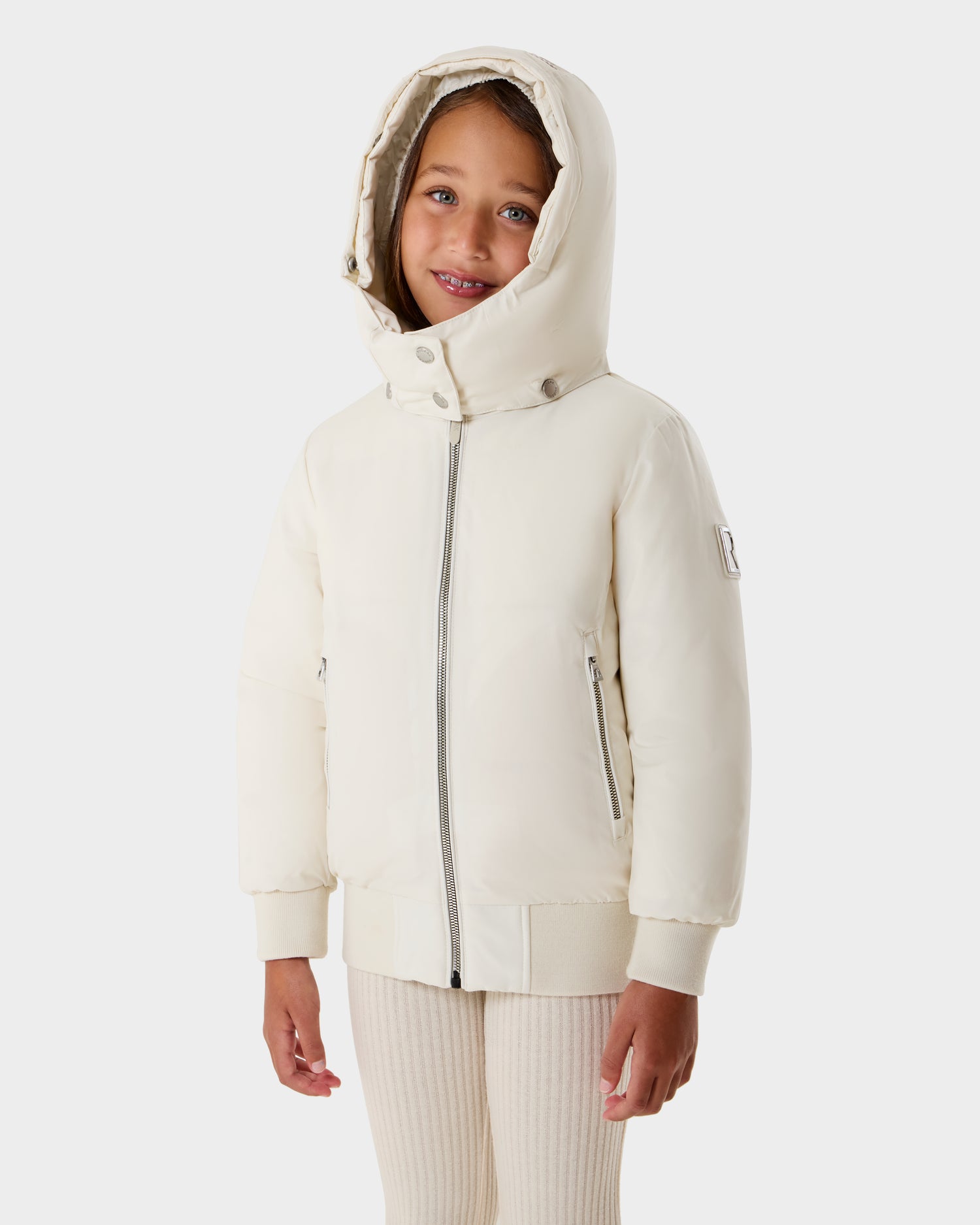 A young girl stands against a white background, smiling slightly while wearing the Rudsak BOOKER K - 0124500 Vanilla kids’ quilted puffer jacket with zipper, ribbed cuffs, and removable hood.