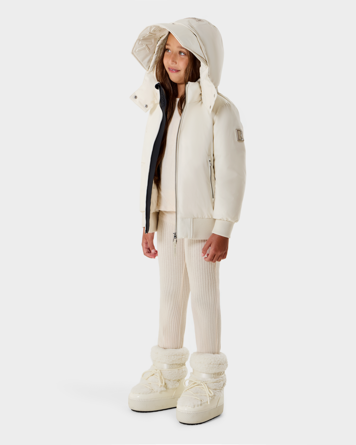A young girl smiles in the Rudsak BOOKER K - 0124500 Vanilla puffer jacket with a removable hood, paired with cream ribbed pants and matching cream winter boots, set against a simple light backdrop.