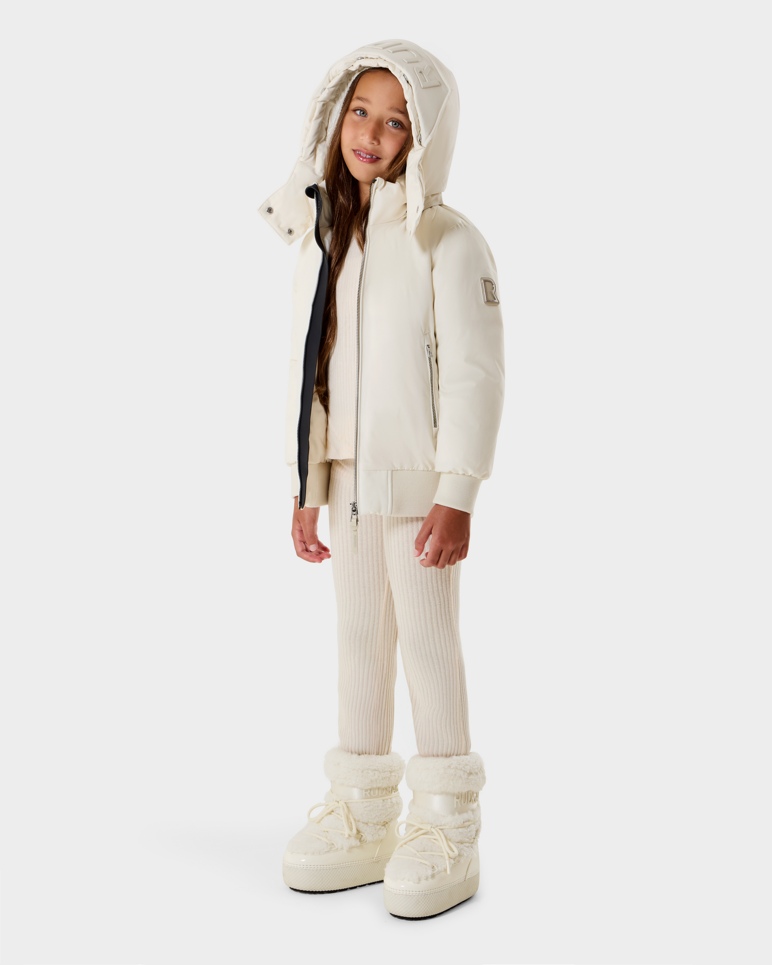 A young girl with long brown hair and a relaxed expression stands against a plain background, wearing the Rudsak BOOKER K - 0124500 Vanilla kids' quilted puffer jacket with removable hood, knit pants, and off-white winter boots.