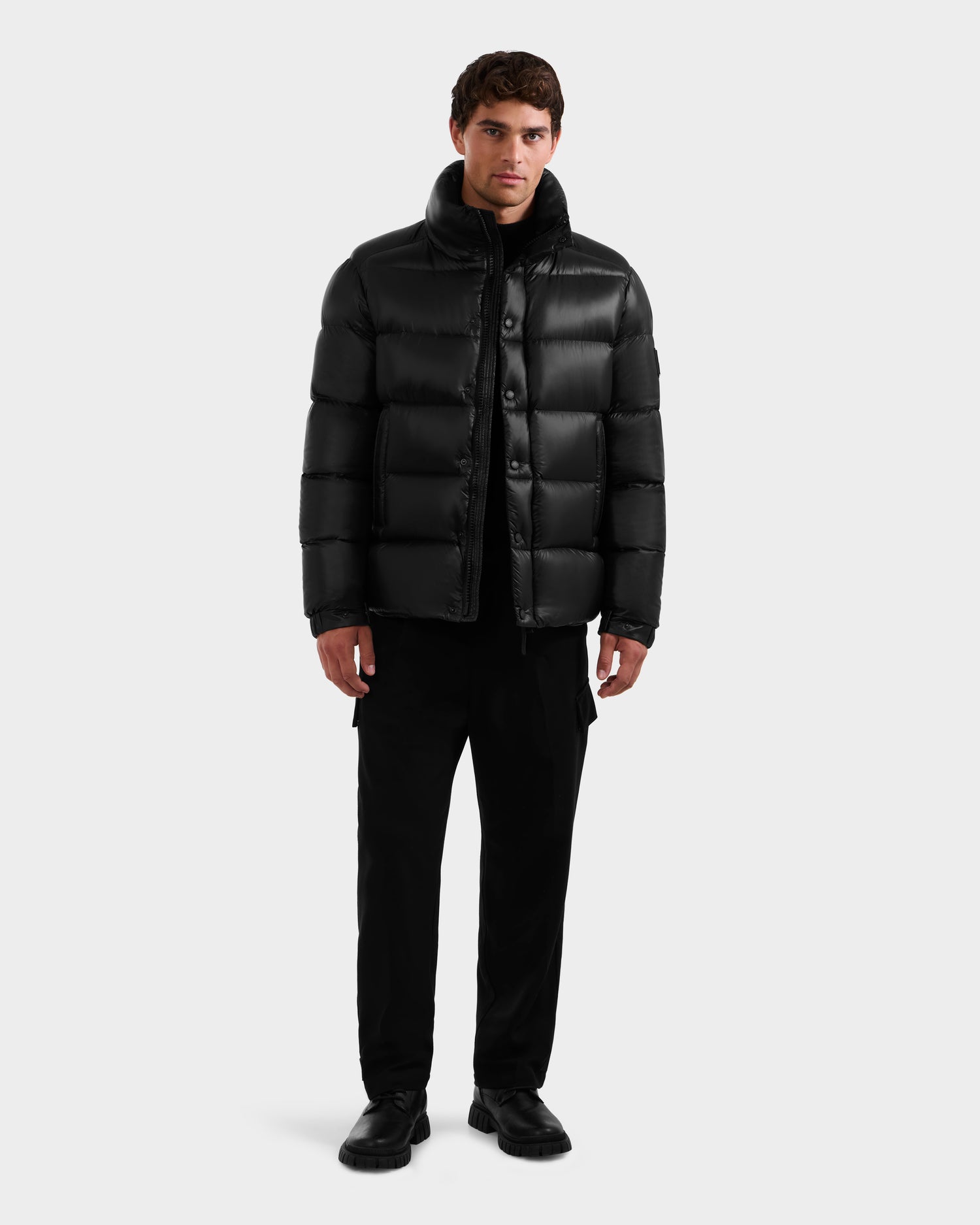 A man stands facing forward wearing the Rudsak BRAYDEN S - M125525 Black quilted down puffer, paired with black pants and black shoes against a plain light gray background.
