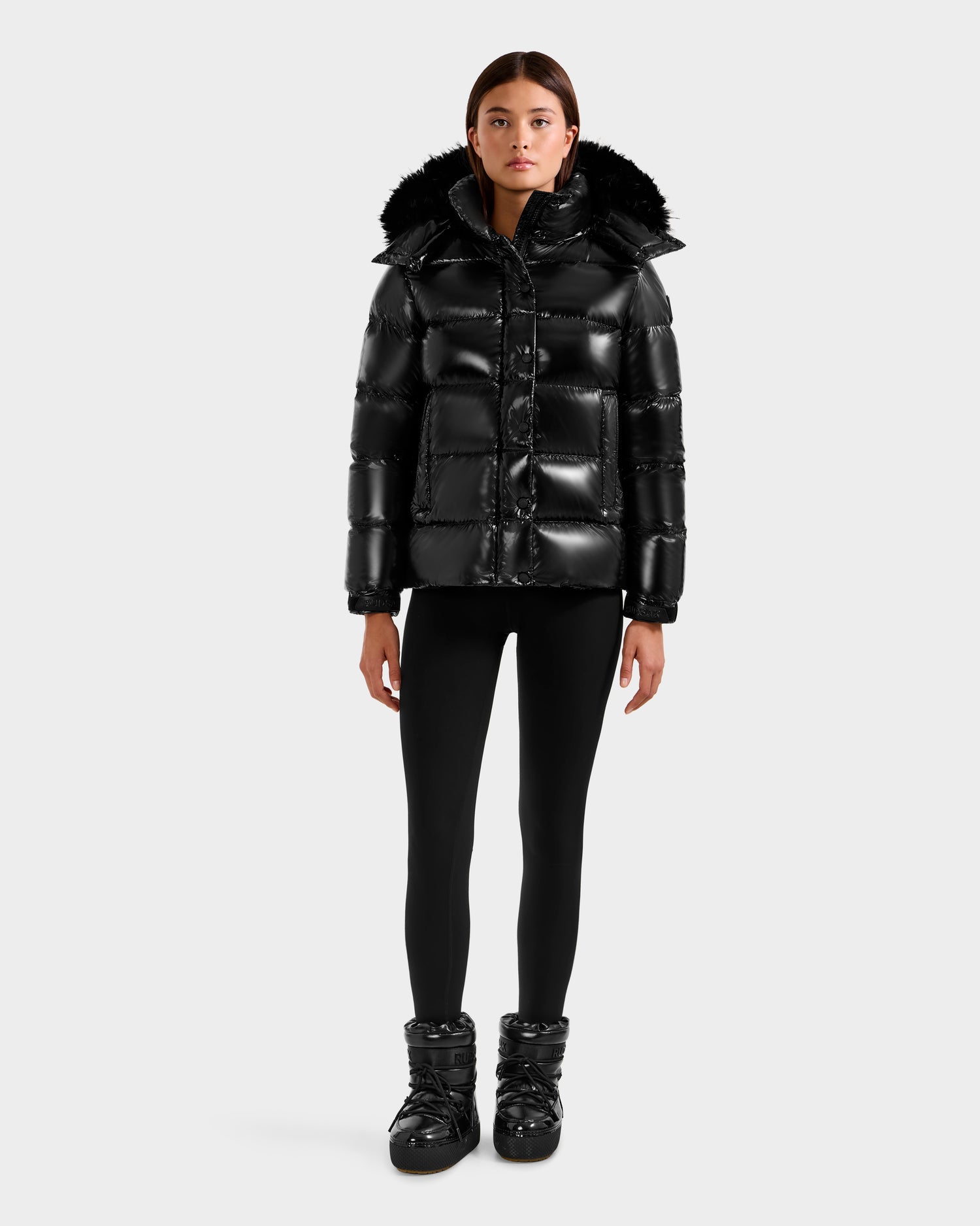 A woman faces forward in a shiny black Rudsak BREE S - L125583 down puffer jacket with a removable fur-lined hood, black leggings, and matching black puffer boots, set against a plain light background.