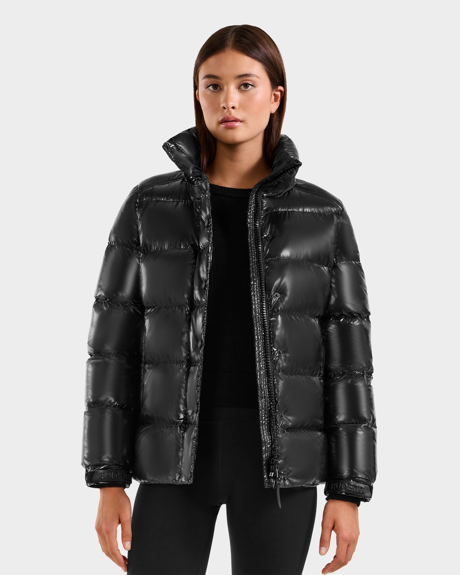 A woman models the Rudsak BREE S - L125583 Black down puffer jacket with a removable hood, styled over a black top and pants, standing against a plain light gray background and looking at the camera.