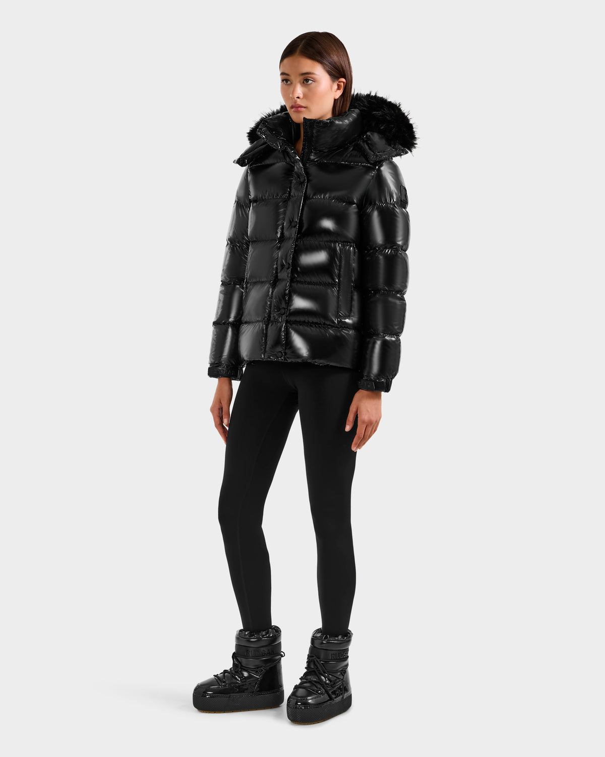 A woman stands against a plain white background wearing Rudsak’s BREE S - L125583 Black down puffer jacket, which features a shiny finish, fur-lined removable hood, black leggings, and winter boots.