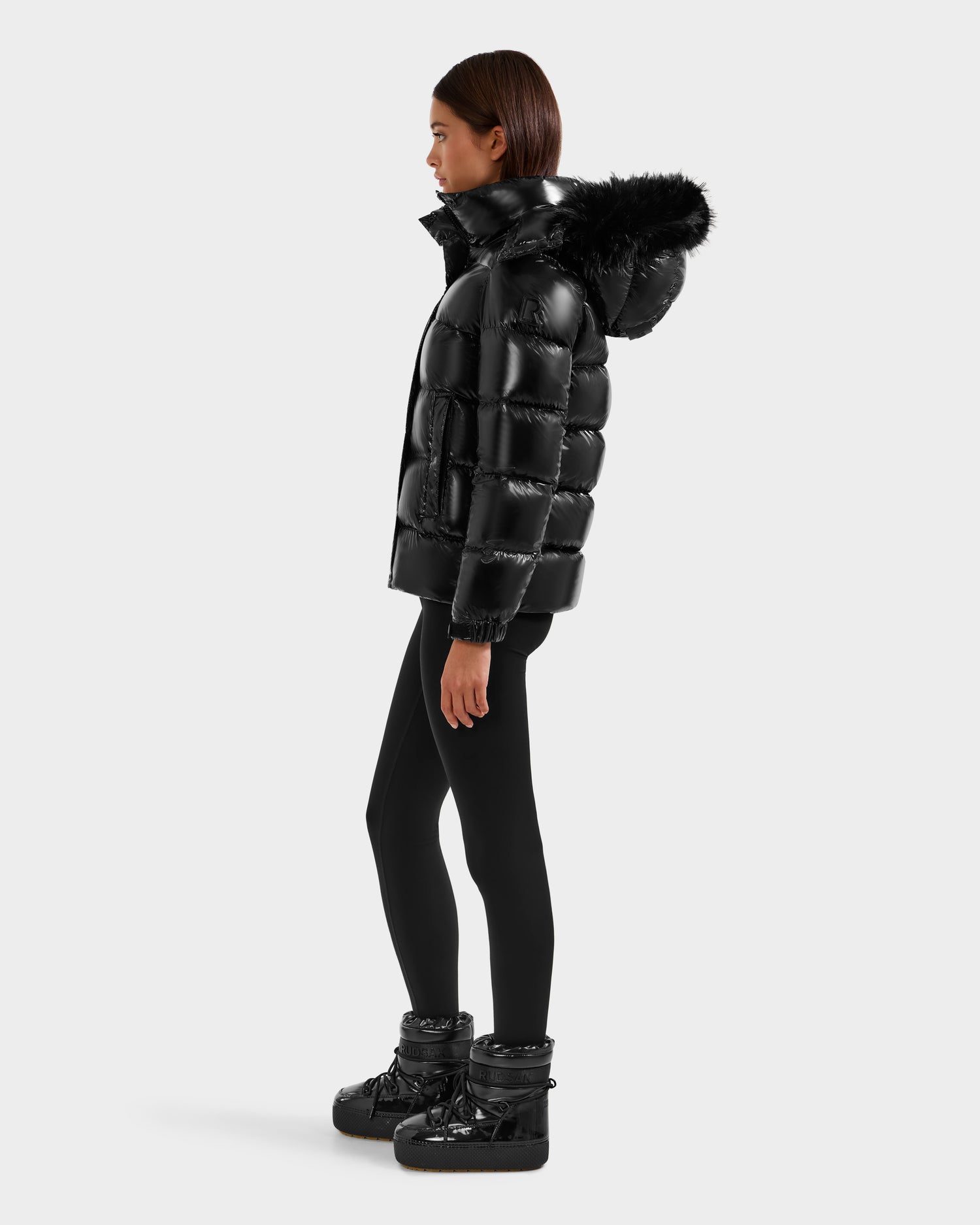 A woman in profile wears the Rudsak BREE S - L125583 Black down puffer jacket with a removable fur-lined hood, paired with black leggings and winter boots, standing against a plain light background.