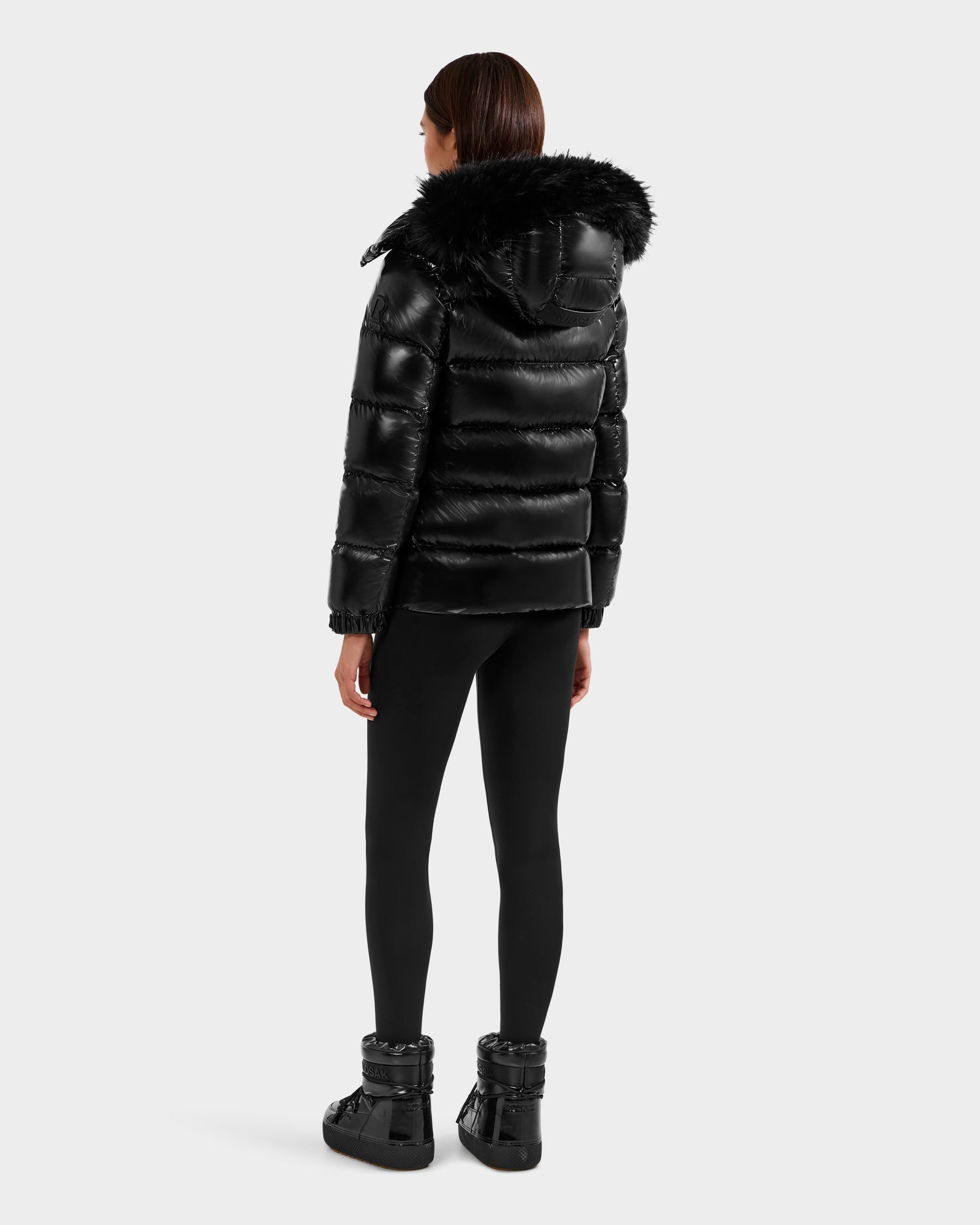 Wearing the Rudsak BREE S - L125583 Black women's down puffer with a weather-resistant, fur-trimmed removable hood, black leggings, and boots, a person stands facing away against a plain light gray background.