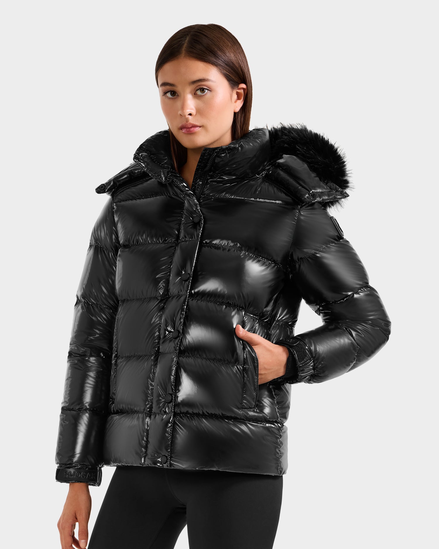 A woman wearing the Rudsak BREE S - L125583 Black down puffer jacket with an ultra-gloss finish and a faux fur-trimmed removable hood stands with one hand in her pocket, looking forward against a plain light background.