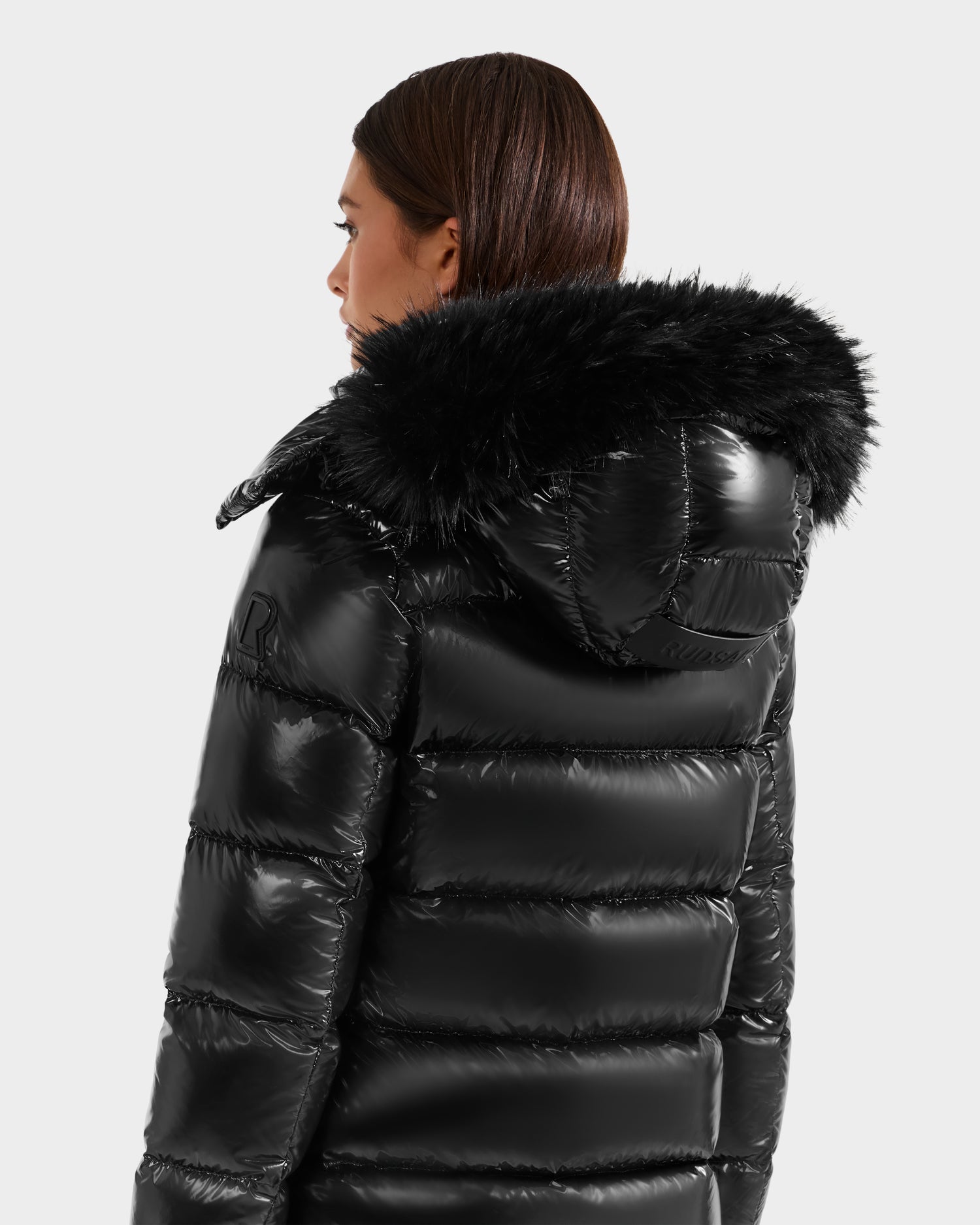 A person with brown hair, seen from behind, wears the Rudsak BREE S - L125583 Black women’s down puffer jacket with a shiny, weather-resistant finish and black faux fur hood, against a plain light gray background.