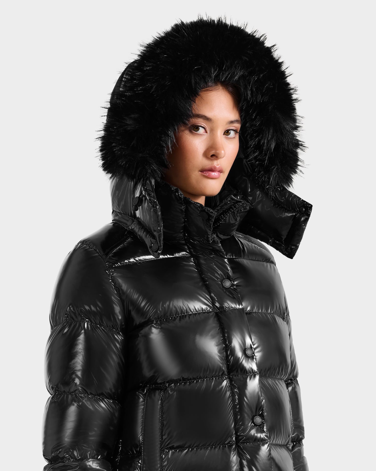 A woman models the Rudsak BREE S - L125583 Black women's down puffer with an ultra-gloss finish and black faux fur–trimmed hood, looking to the side against a plain light gray background.