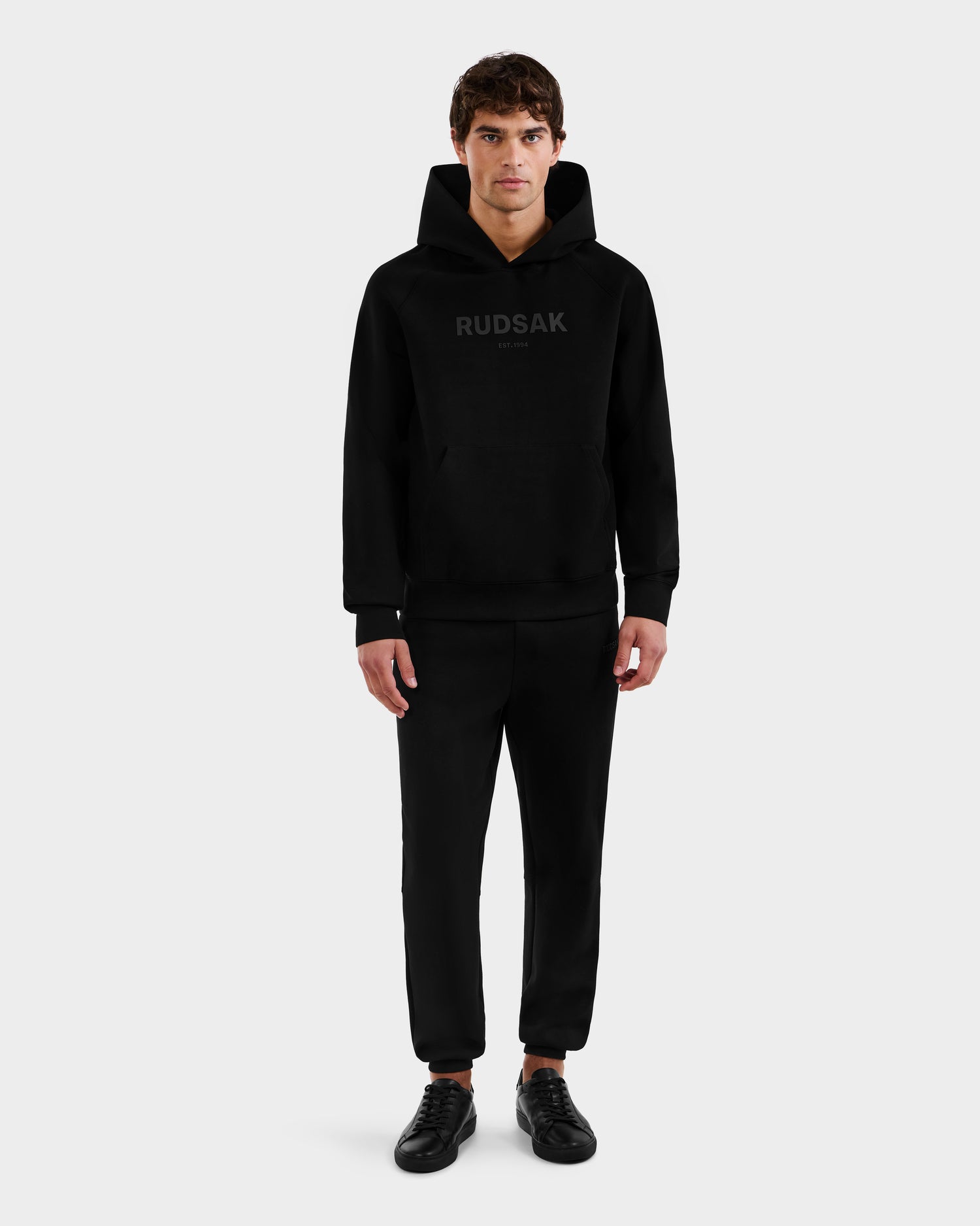 A man stands facing forward against a plain light gray background, wearing the Rudsak BRIAR - 9525001 Black M hoodie, paired with black pants and black sneakers.