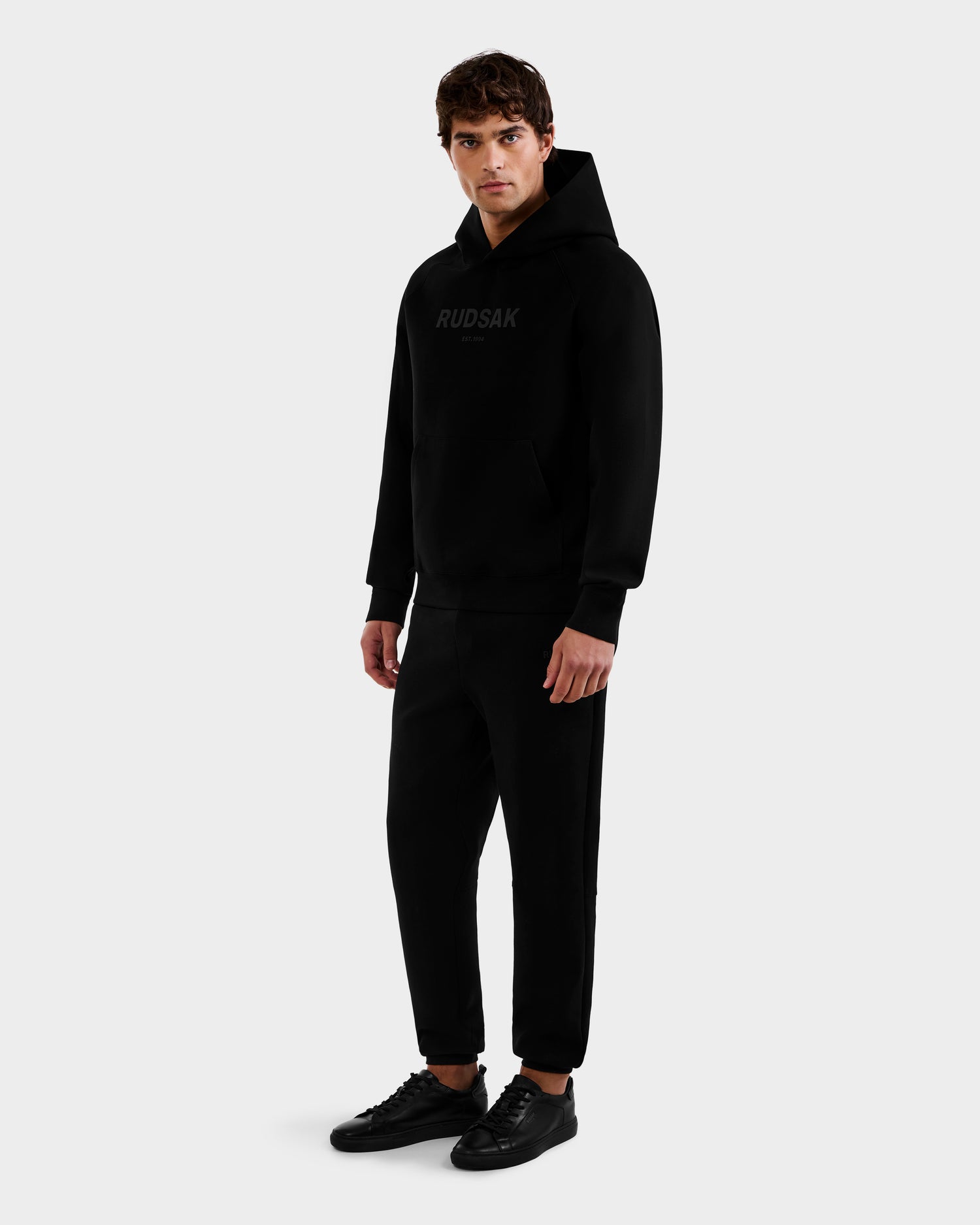 A young man stands against a plain background, wearing the Rudsak BRIAR - 9525001 Black M premium cotton hoodie with matching jogger pants and black sneakers. He faces forward with a neutral expression and hands at his sides.