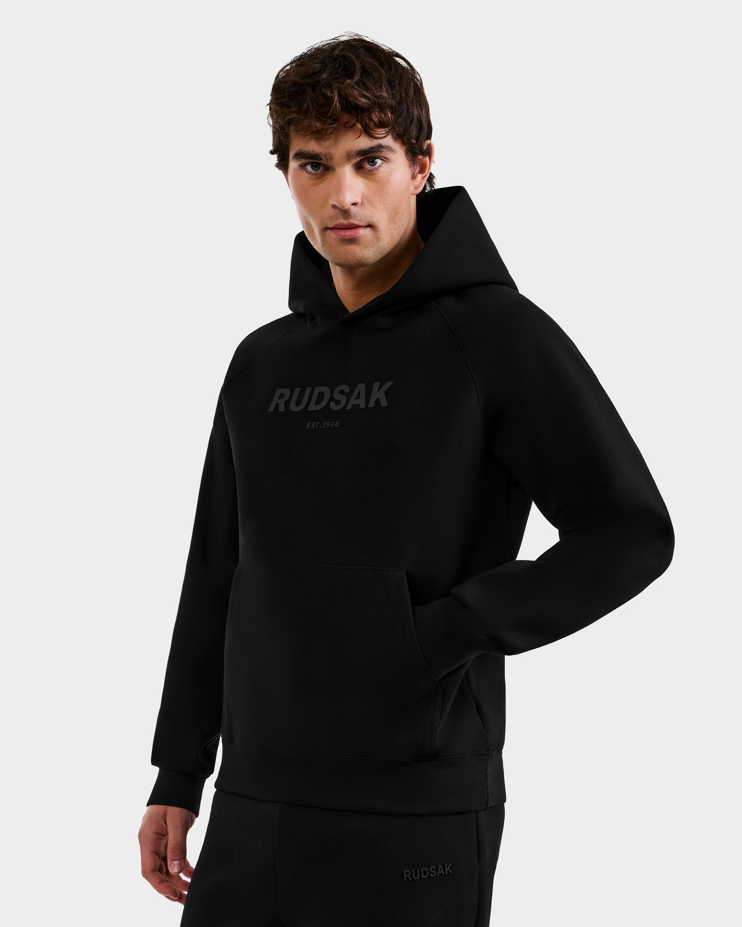 Someone wearing the Rudsak BRIAR - 9525001 Black M unisex hoodie, featuring a front logo and premium cotton blend, stands with one hand in the pocket against a plain light background, showcasing its casual style.