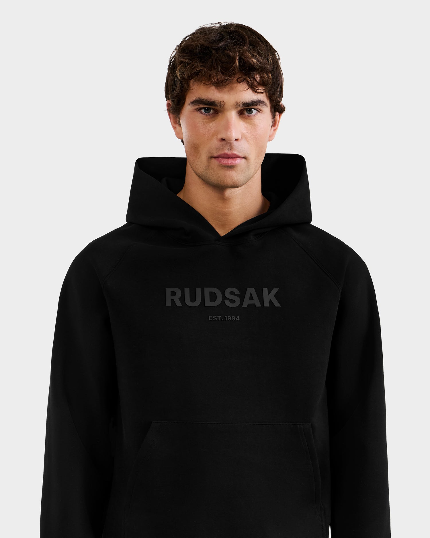 A young man with short brown hair wears the Rudsak BRIAR - 9525001 Black M premium cotton hoodie, featuring subtle dark "RUDSAK EST. 1994" print on the front, standing against a plain light grey background.