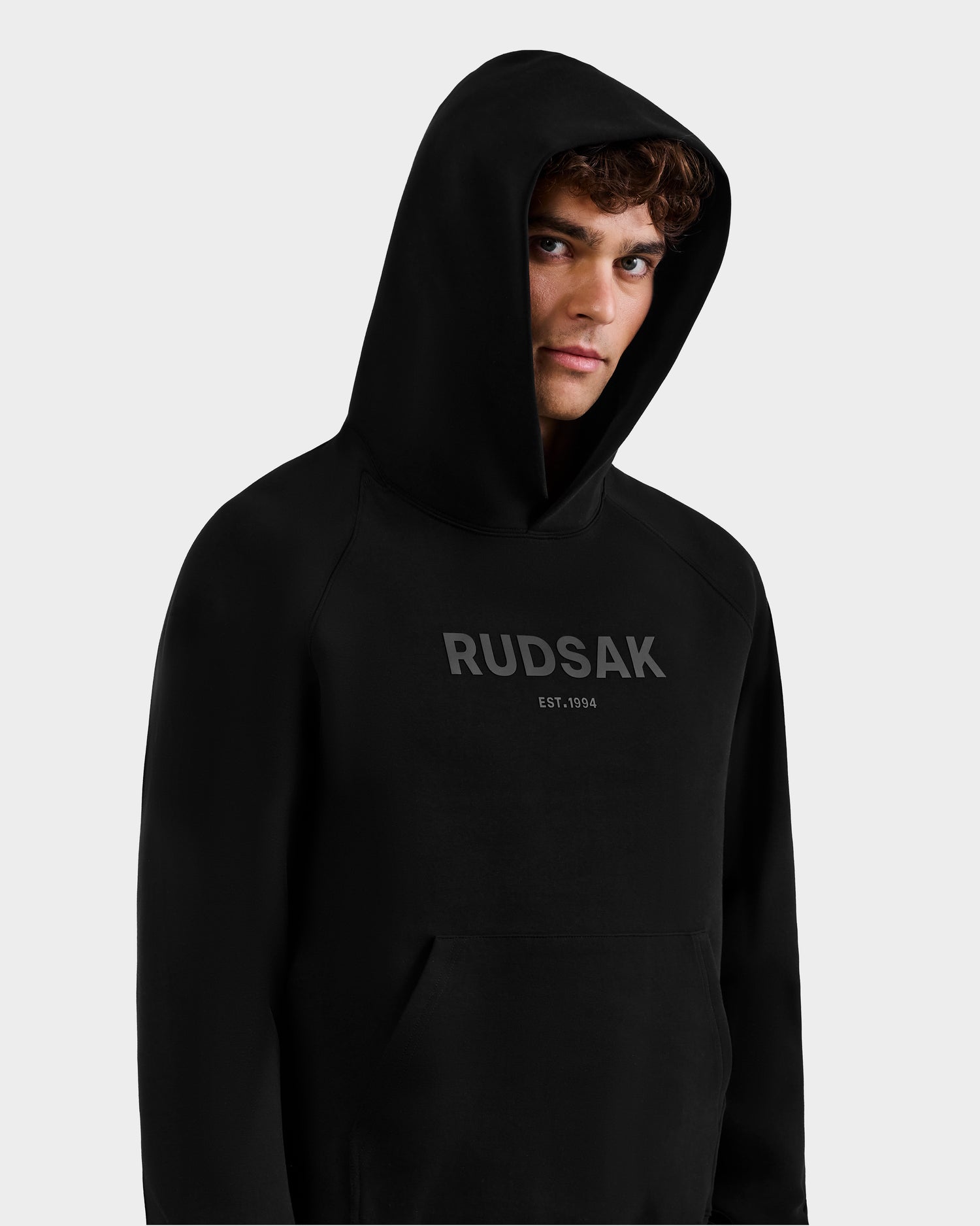 A person wearing the Rudsak BRIAR - 9525001 Black M unisex hoodie with the hood up and "RUDSAK EST. 1994" in gray on the front stands against a plain light background, highlighting this premium cotton hoodie’s sleek style.