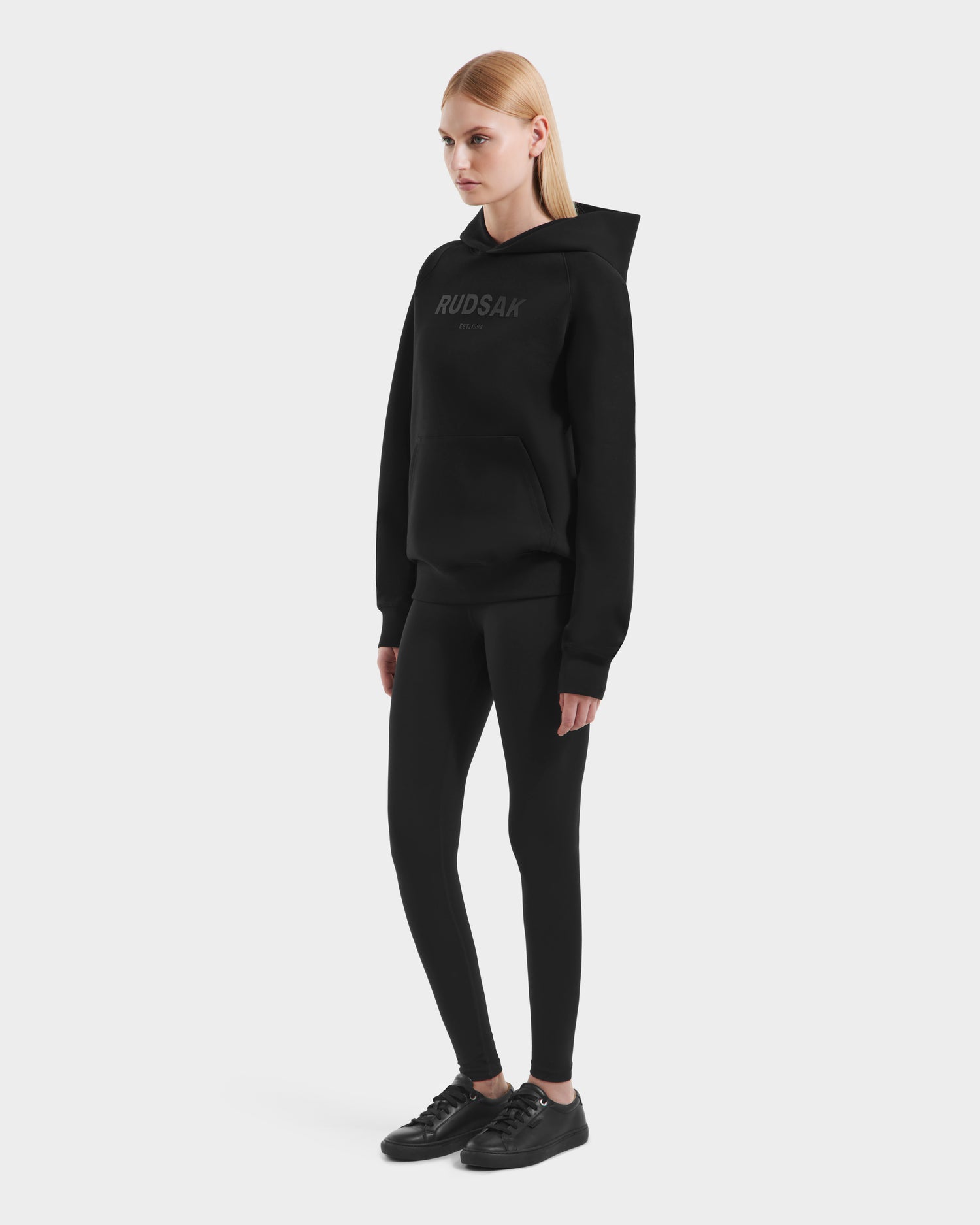 A woman with straight blonde hair wears the Rudsak BRIAR - 9525001 Black W unisex hoodie, styled with black leggings and sneakers, set against a plain white background.