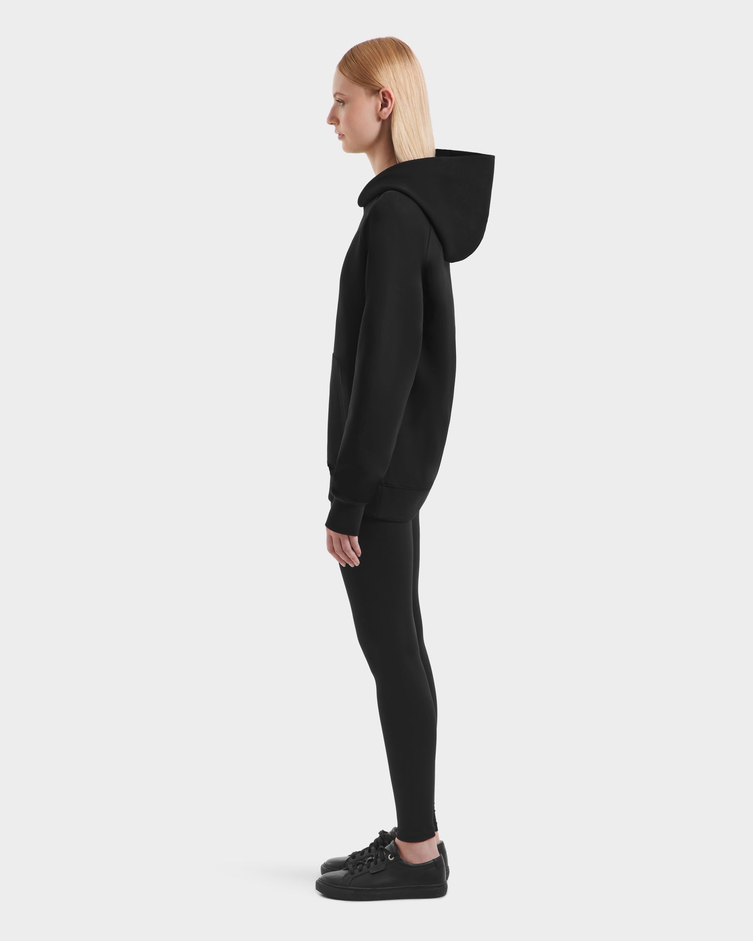 Side view of a person with straight blonde hair wearing the Rudsak BRIAR - 9525001 Black W unisex hoodie, black leggings, and black sneakers on a white background. The hoodie is crafted from a premium cotton blend for comfort.