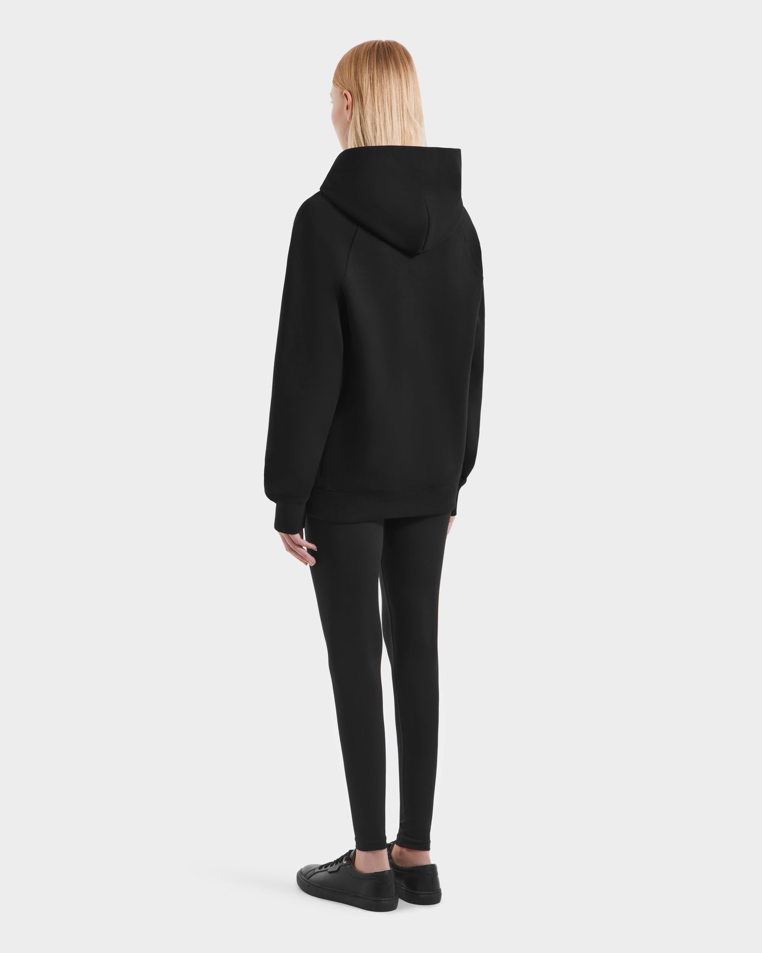 A person with straight, blonde hair faces away, wearing the Rudsak BRIAR - 9525001 Black W hoodie with black leggings and shoes against a plain white background.