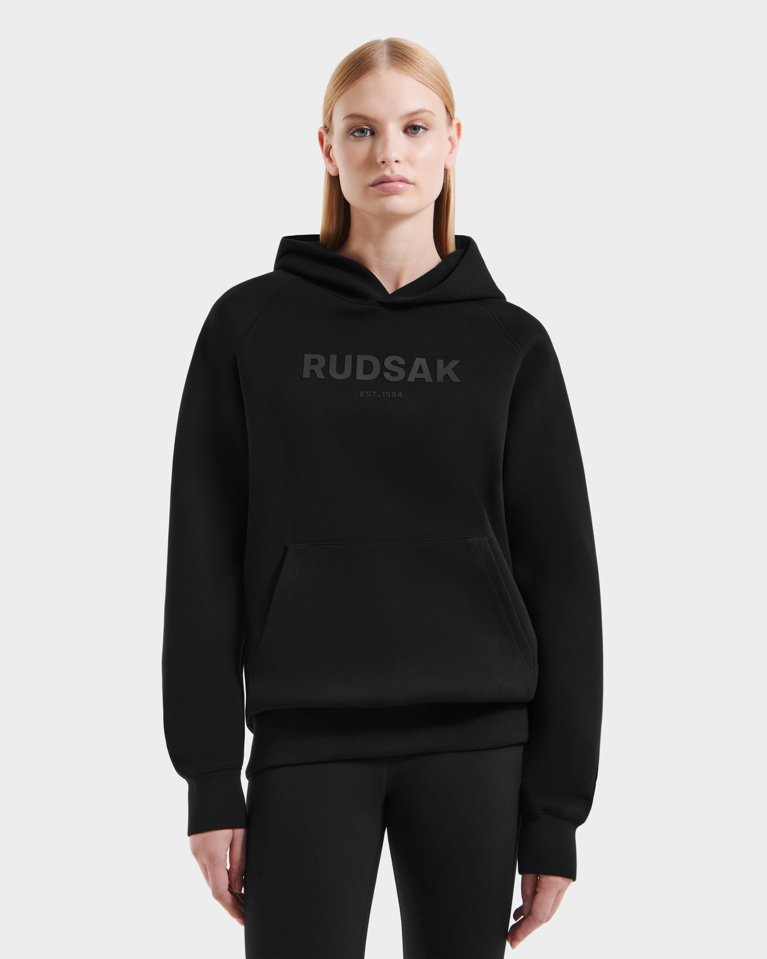 A woman with straight blonde hair wears the Rudsak BRIAR - 9525001 Black W unisex hoodie, made from a premium cotton blend with "RUDSAK EST. 1994" on the front, paired with black pants against a plain white background for a casual look.