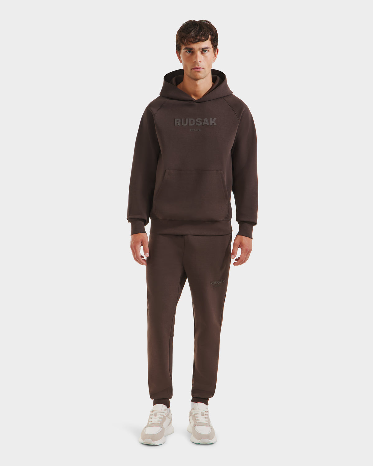 A man faces forward in a casual look, wearing the Rudsak BRIAR - 9525001 Espresso M unisex hoodie and sweatpants set in premium cotton blend with RUDSAK on the hoodie, paired with white sneakers against a plain white background.