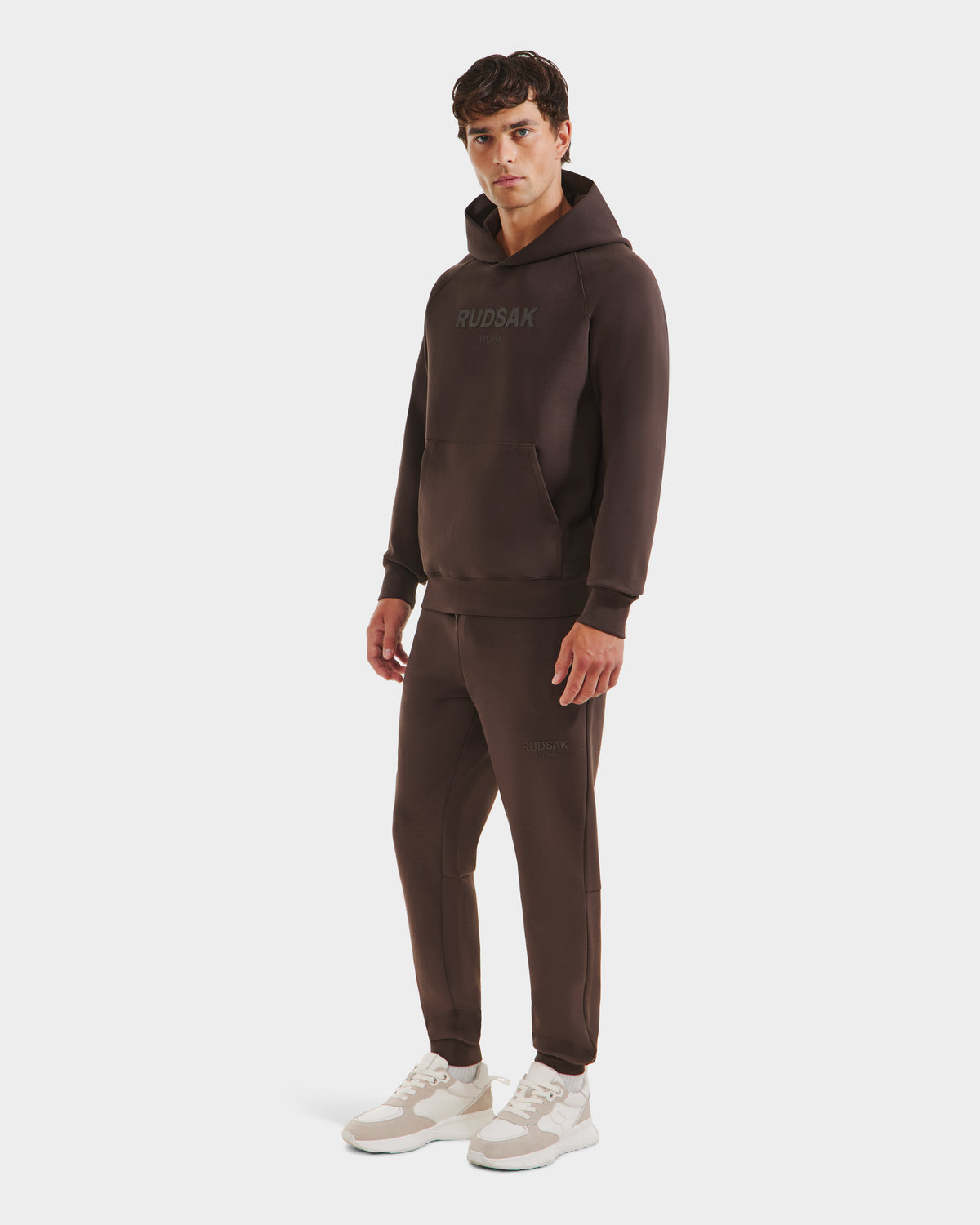 A man wears the Rudsak BRIAR - 9525001 Espresso M dark brown unisex hoodie and joggers, made from premium cotton blend with branding, paired with white sneakers for a relaxed, casual look against a plain white background.