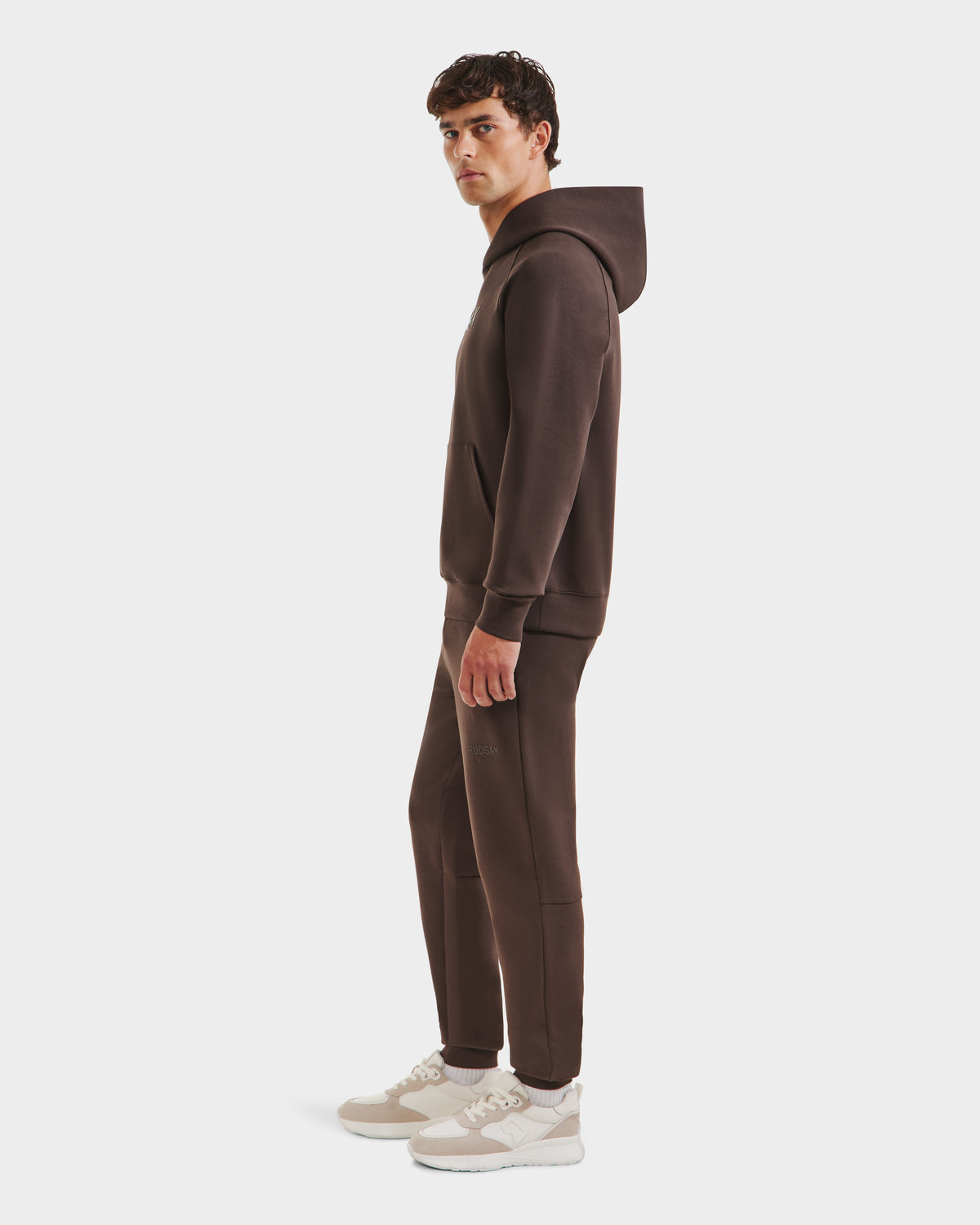 A man stands sideways in the Rudsak BRIAR - 9525001 Espresso M unisex hoodie and joggers, paired with beige and white sneakers, creating a modern casual look against a plain white background.