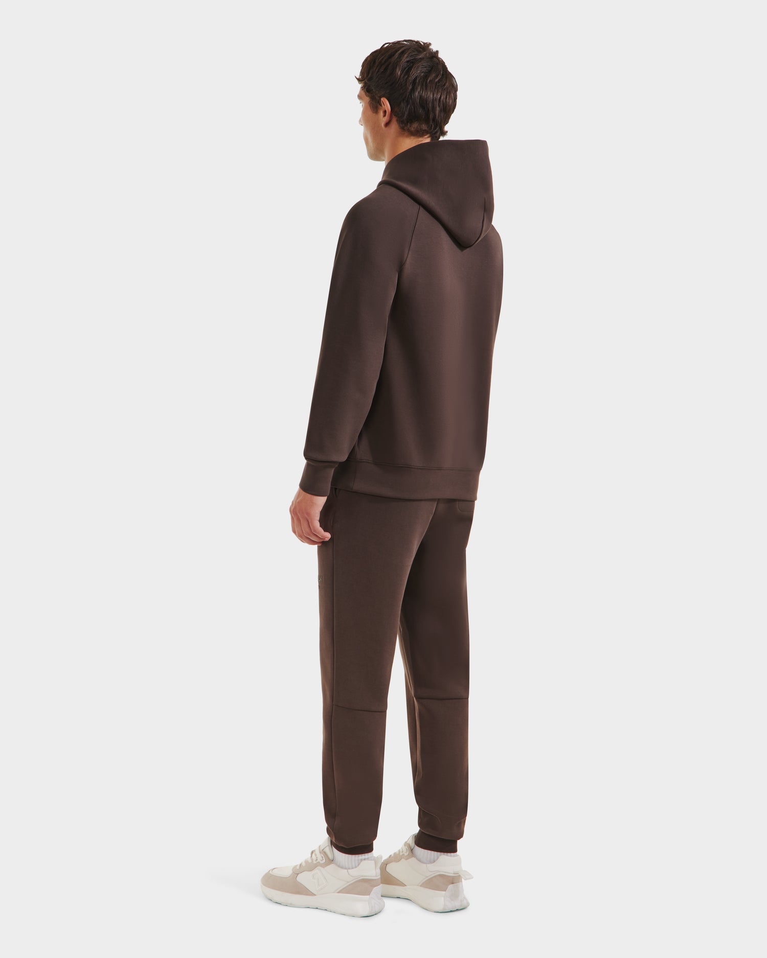 A man faces away, wearing the Rudsak BRIAR - 9525001 Espresso M unisex hoodie and matching brown sweatpants, made from a premium cotton blend with white sneakers, set against a plain white background.