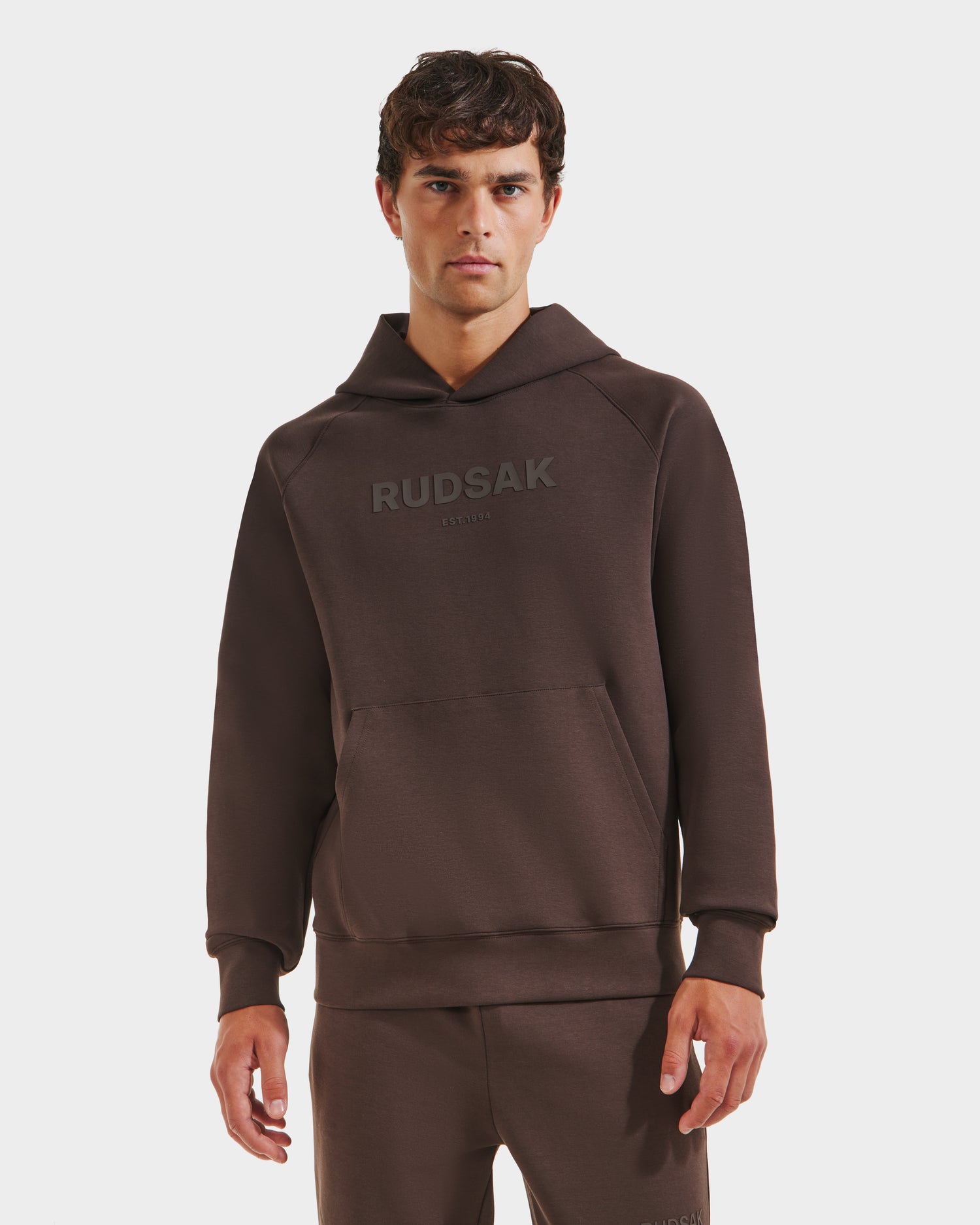 A man with short brown hair and a neutral expression stands facing forward, wearing the Rudsak BRIAR - 9525001 Espresso M premium cotton hoodie with branding on the chest. The background is plain white.