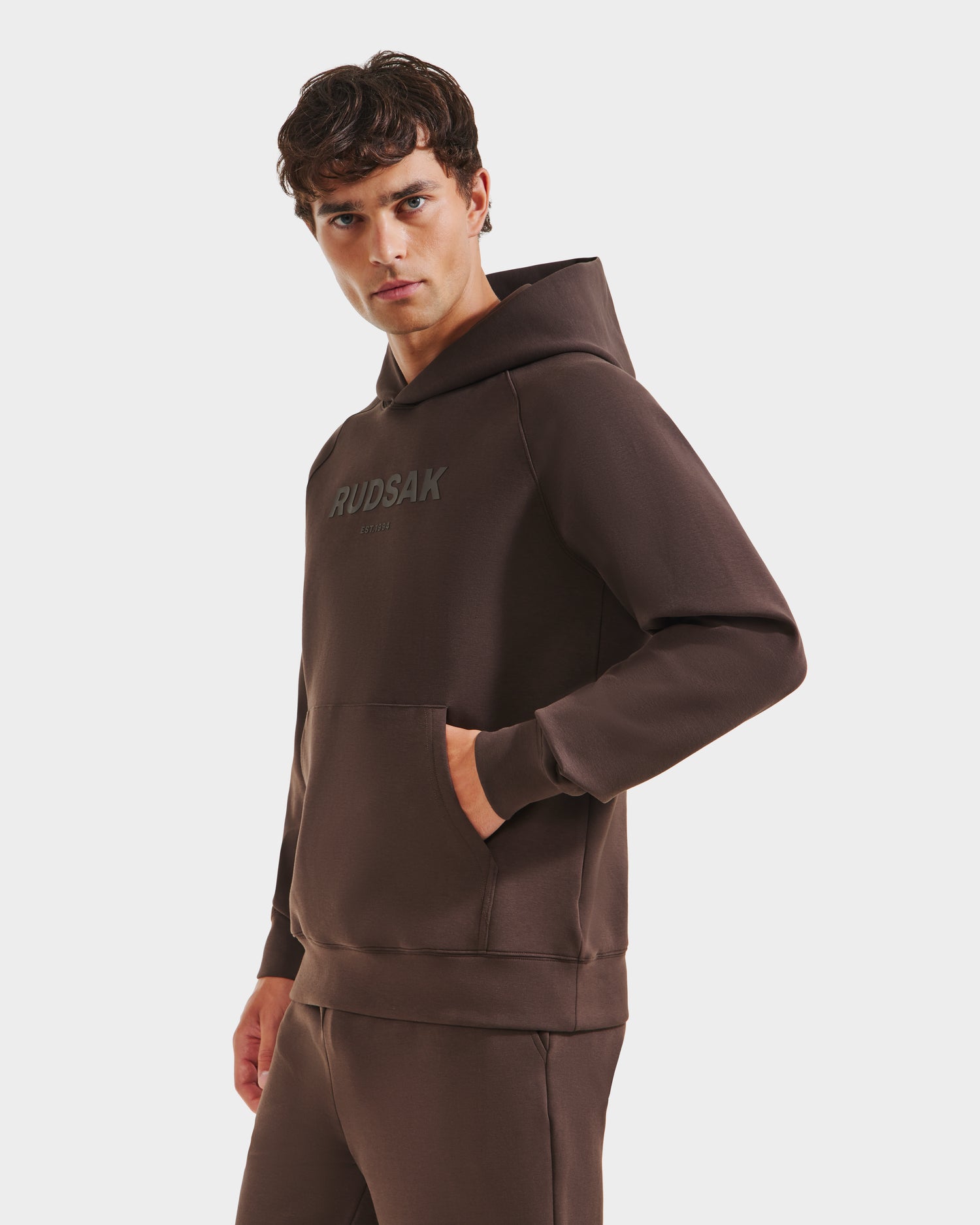A man wears the Rudsak BRIAR - 9525001 Espresso M unisex brown hoodie and pants, made from a premium cotton blend with RUDSAK on the chest, standing against a plain white background and looking forward.