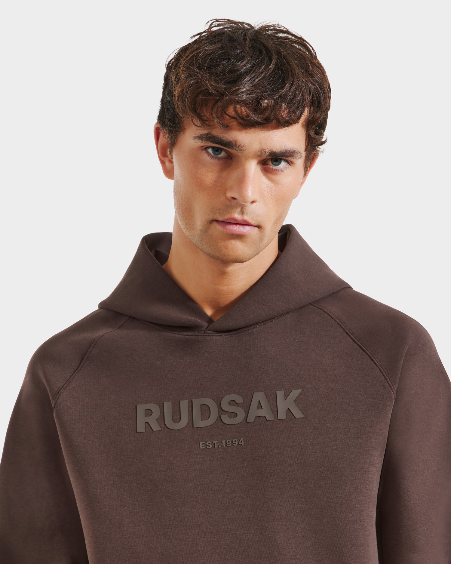 A young man with short, curly brown hair models the Rudsak BRIAR - 9525001 Espresso M unisex hoodie in premium cotton blend with RUDSAK EST. 1994 on the chest, standing against a plain white background for a casual look.