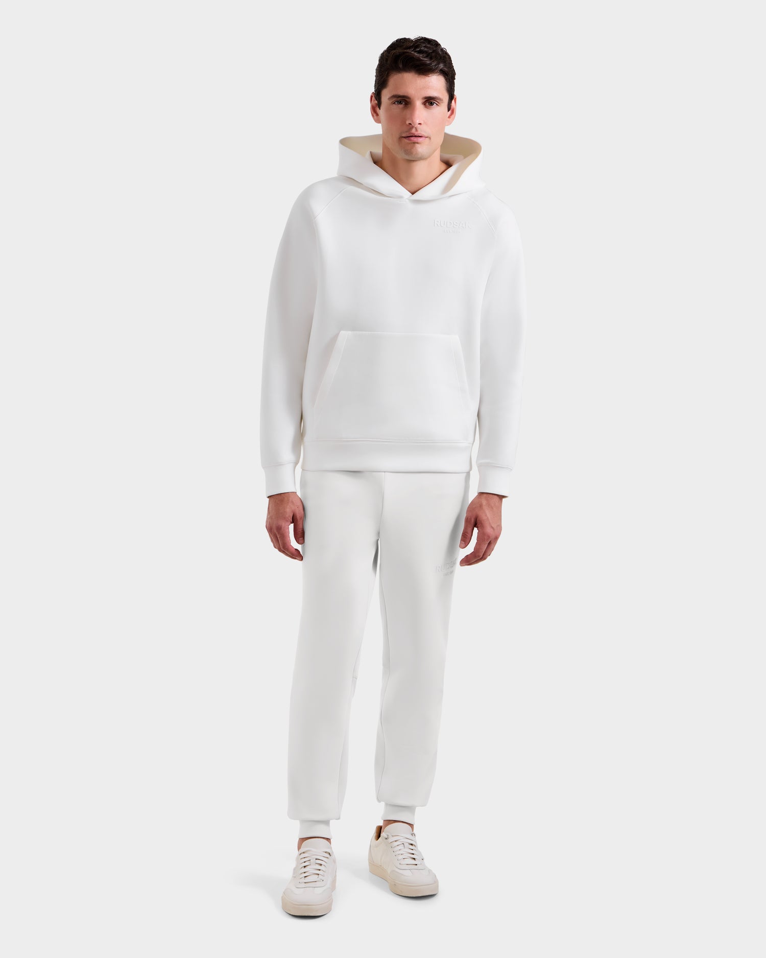 A man faces forward wearing the Rudsak BRIAR - 9525001 Iceland M unisex hoodie with a front pocket, paired with matching white jogger pants and white sneakers, set against a plain light gray background.