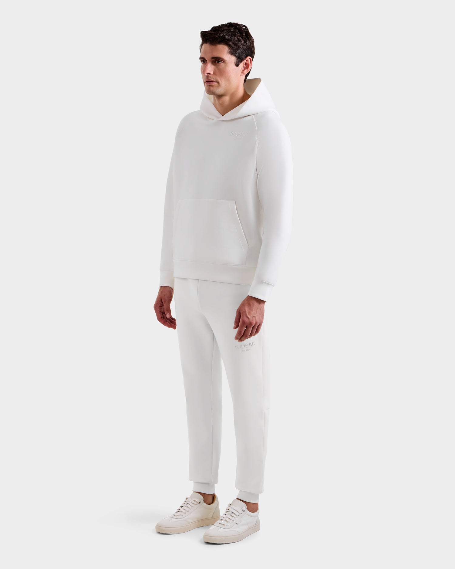 A man stands against a plain white background, wearing the Rudsak BRIAR - 9525001 Iceland M unisex hoodie and jogger pants with sneakers for a casual look.
