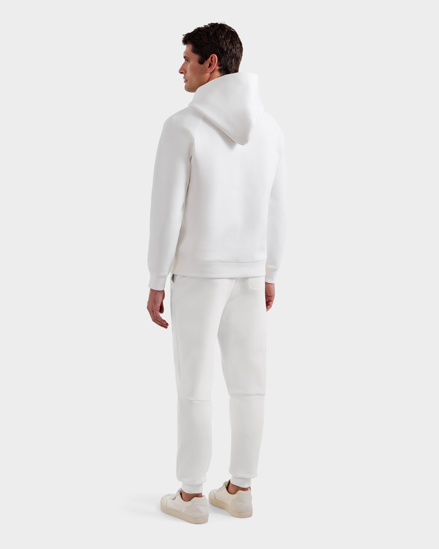A man stands facing away, wearing Rudsak’s BRIAR - 9525001 Iceland M unisex hoodie, white sweatpants, and sneakers—crafted from a premium cotton blend—against a plain light background for a modern casual look.