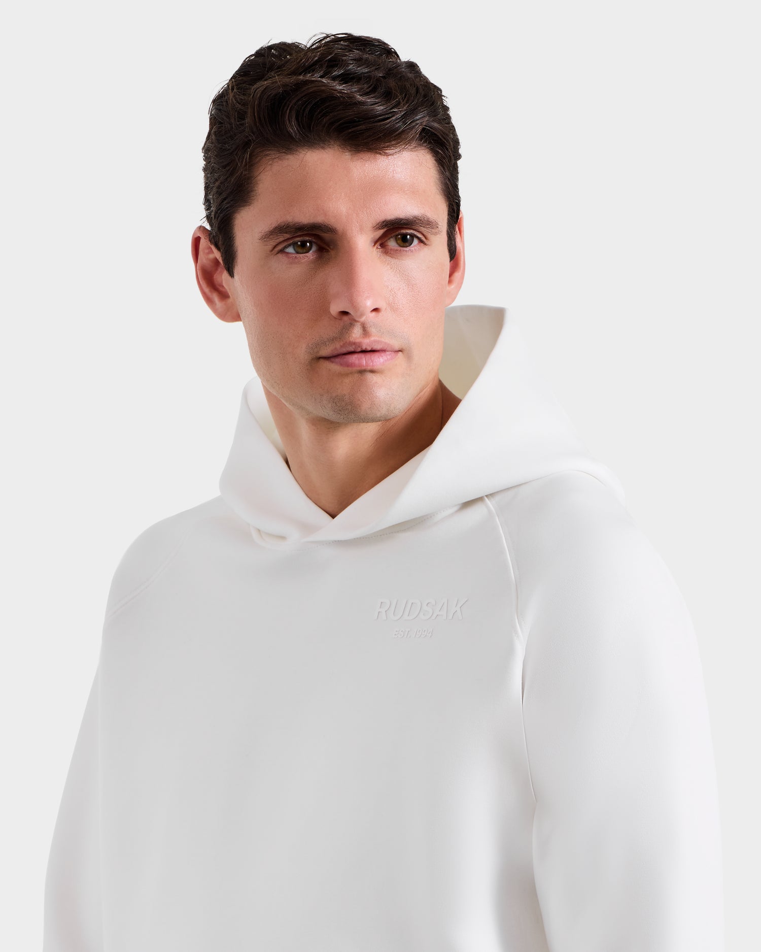 A man models the Rudsak BRIAR - 9525001 Iceland M unisex white hoodie with RUDSAK embossed on the chest, gazing sideways against a light background to showcase its casual, premium cotton blend style.