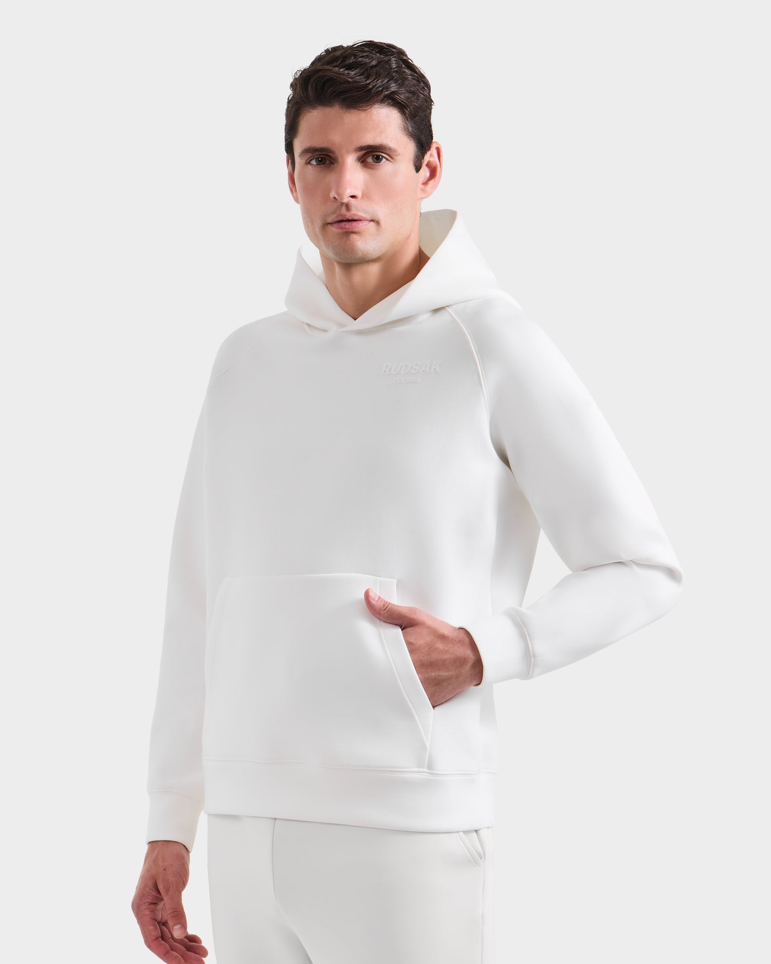 A man with short brown hair models the Rudsak BRIAR - 9525001 Iceland M hoodie and matching white pants, standing against a plain light gray background with one hand in his pocket for a relaxed, casual look.
