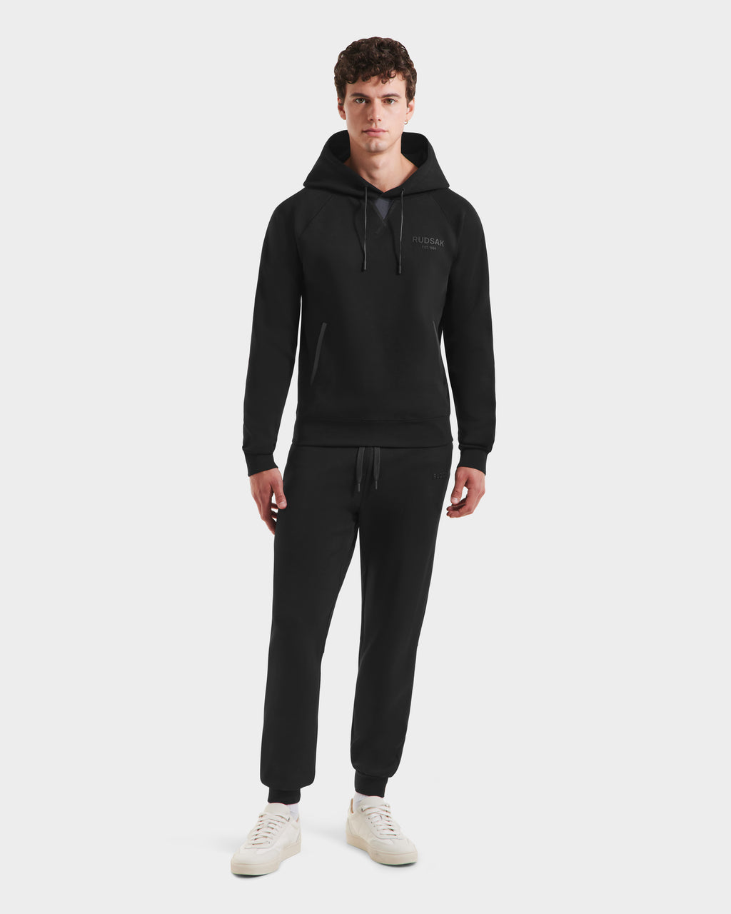 A young man faces forward, modeling Rudsak’s BROOKLYN - 6525593 Black hoodie and jogger set with white sneakers, set against a plain light background.